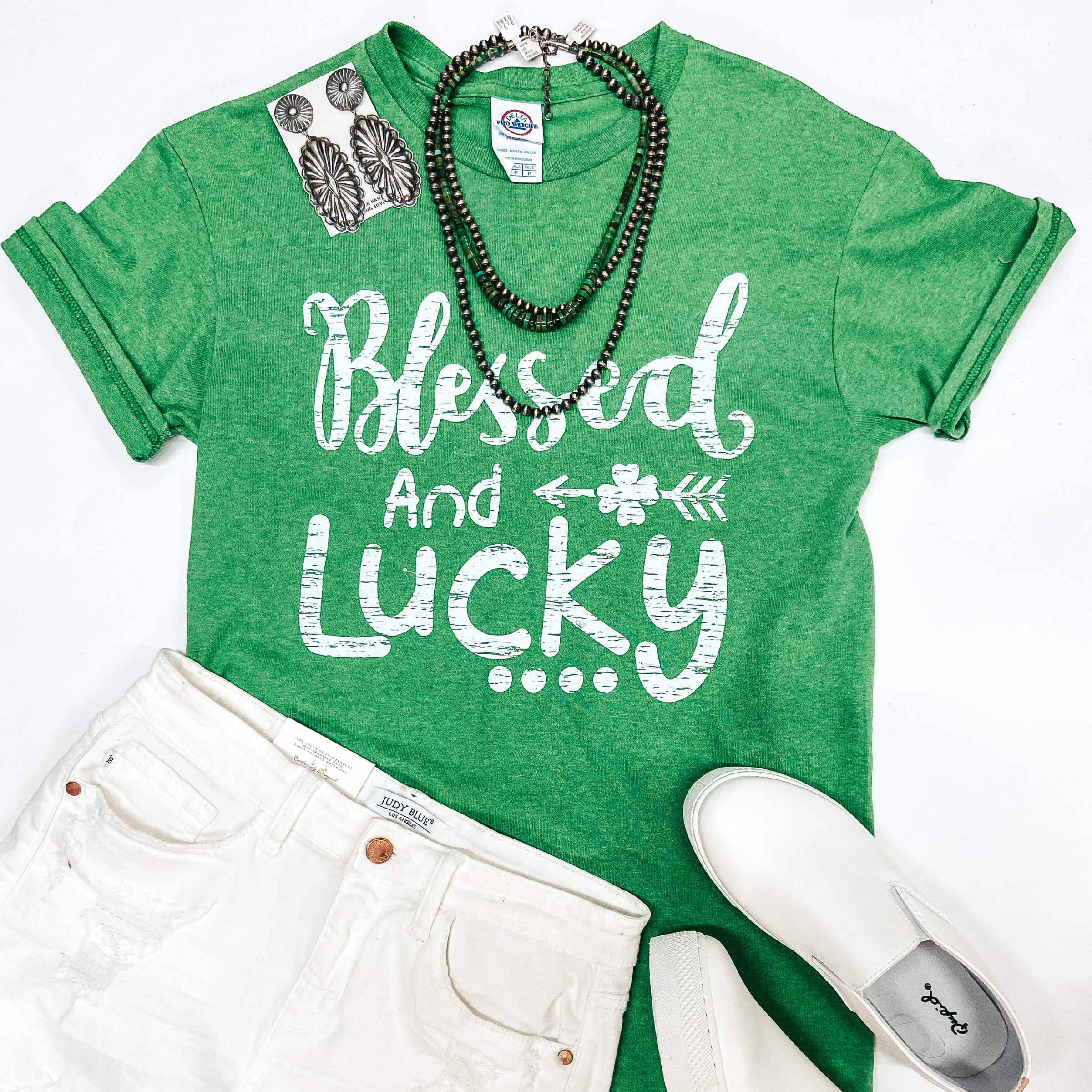 A green graphic tee that says "Blessed and Lucky" with an arrow and clover. Pictured with white shorts, sneakers, and silver jewelry.