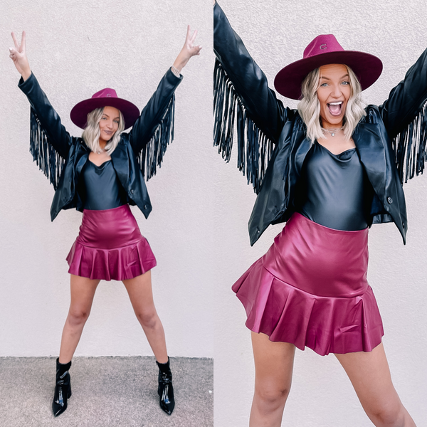 Model is wearing a faux, leather mini skort in maroon. Model has it paired with a black leather bodysuit, black booties, a maroon hat, and a black fringe jacket. 