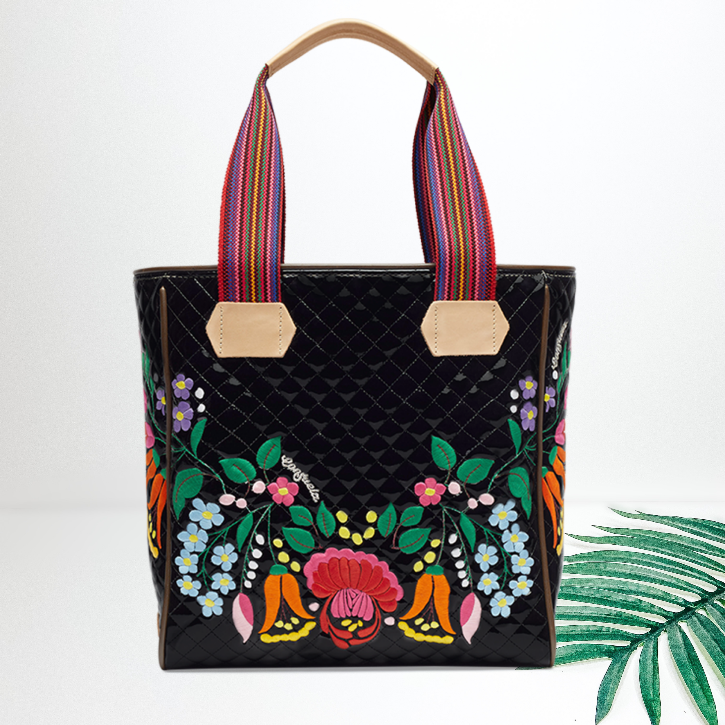 A black quilted tote bag with floral embroidery on the front. Pictured on white background with a palm leaf.