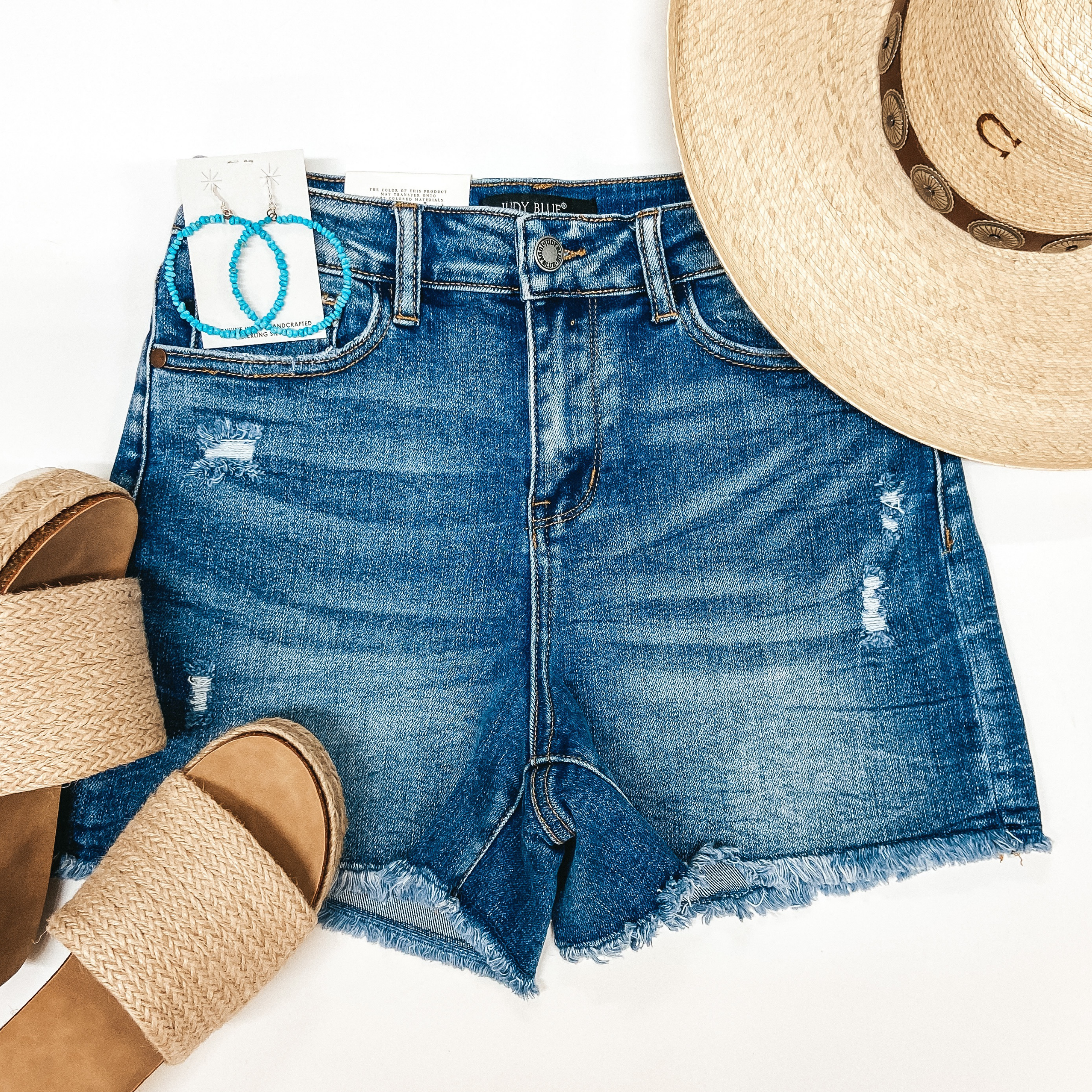 A pair of dark wash cut off shorts. Pictured on white background with straw hat, turquoise earrings, and a pair of platform sandals.