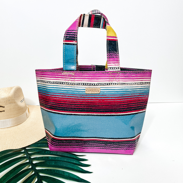 A small size serape print bag that is a mix of blue, yellow, and purple. Pictured on white background with a palm leaf and straw hat.
