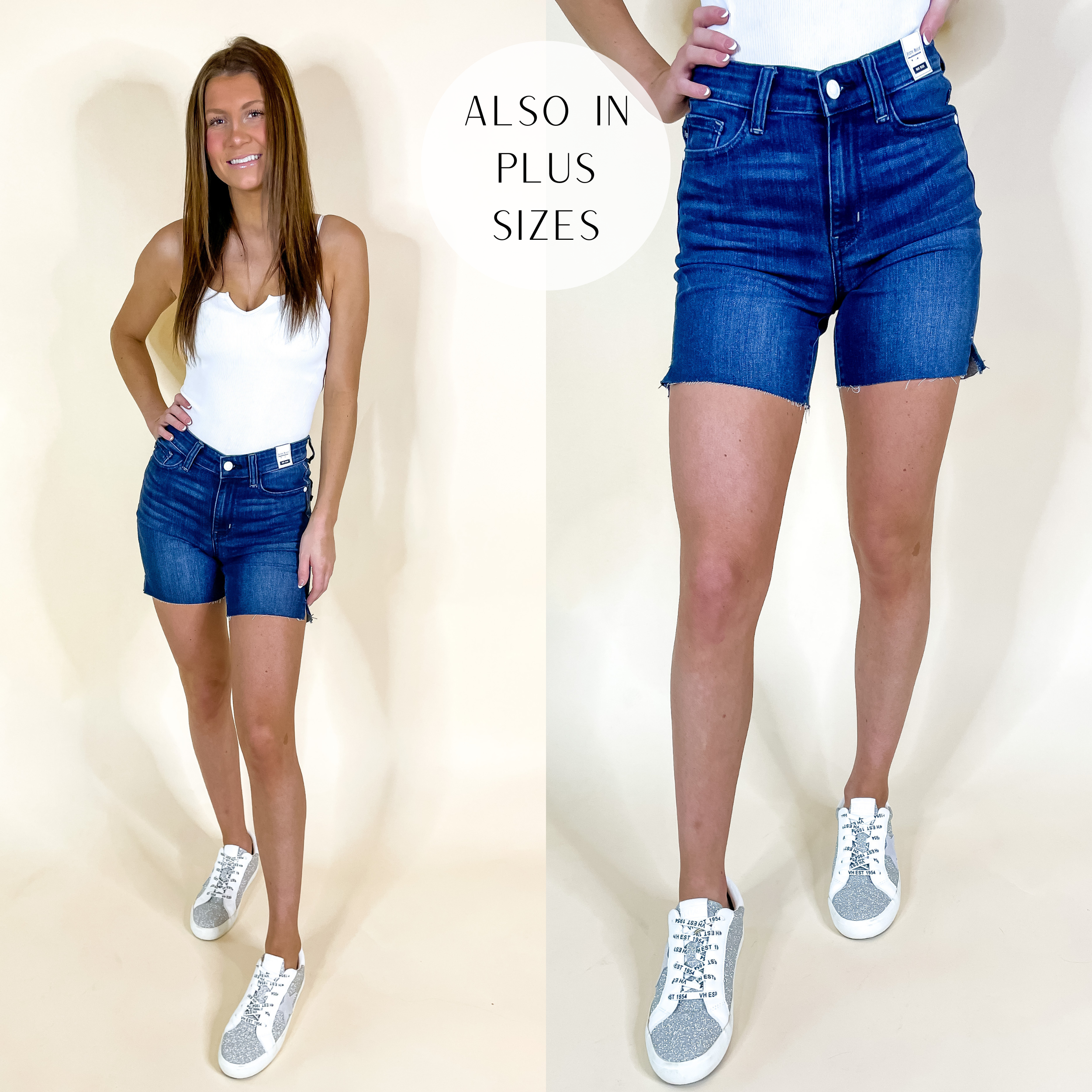Model is wearing a pair of dark wash denim shorts with a raw hem and a mid-thigh length. Model has these shorts paired with white sneakers and a white tank top.