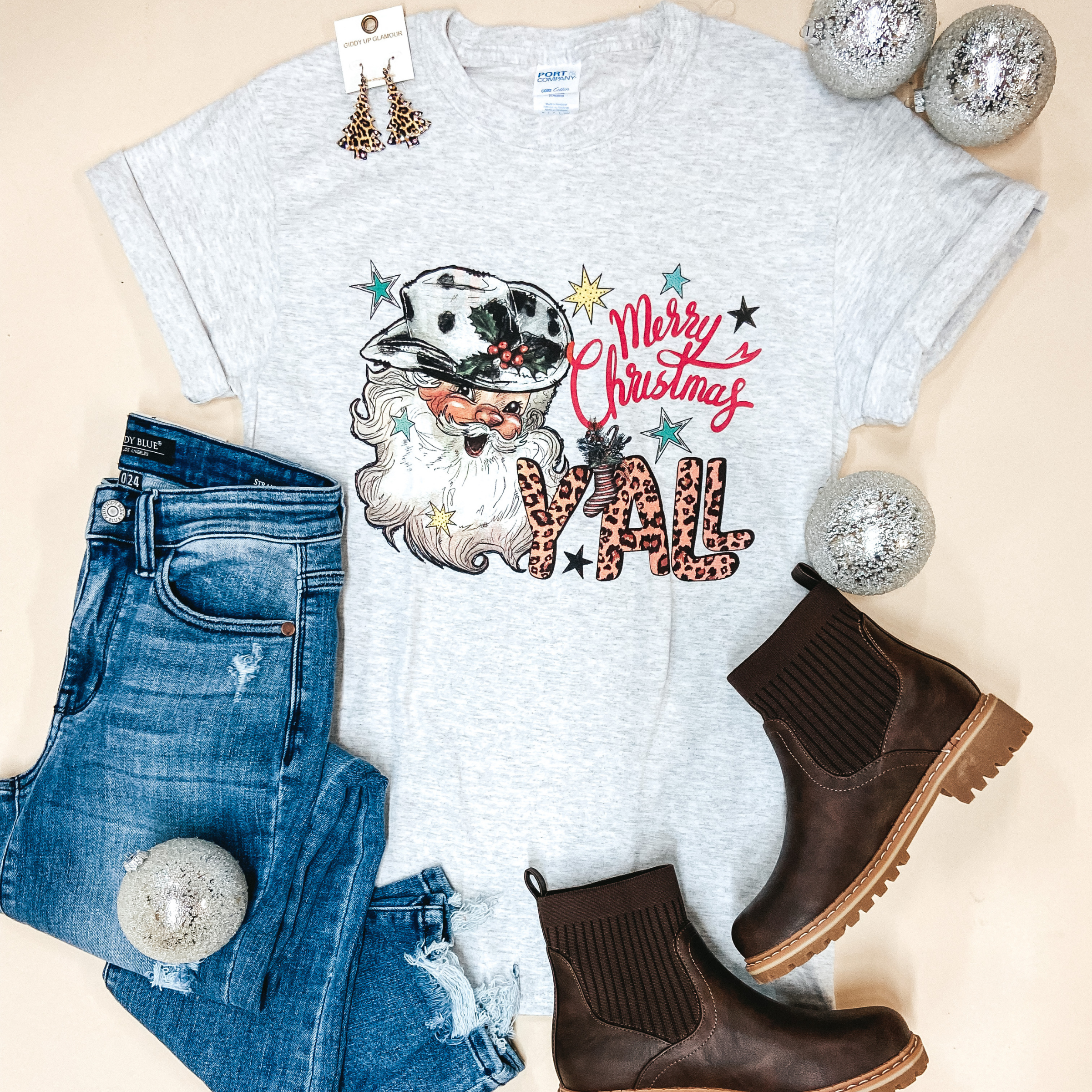 A grey short sleeve graphic tee with a crew neckline and Western Santa Clause graphic that says "Merry Christmas Y'all." Pictured on a white background with distressed jeans, brown booties, and leopard print Christmas Tree earrings.