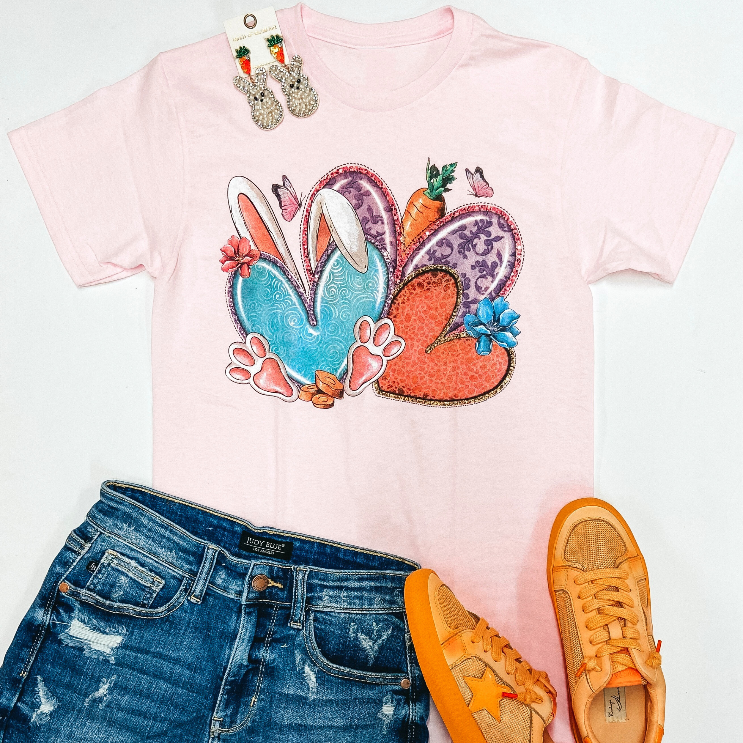 A light pink tee shirt with short sleeves and a graphic of colorful hearts, butterflies, carrots, bunny ears and feet. This tee is pictured on a white background with orange sneakers, peep shaped earrings, and denim shorts.
