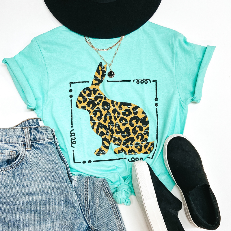 A mint colored tee shirt with a leopard print bunny rabbit graphic. Pictured on white background with black sneakers, distressed jeans, a black hat, and gold jewelry.
