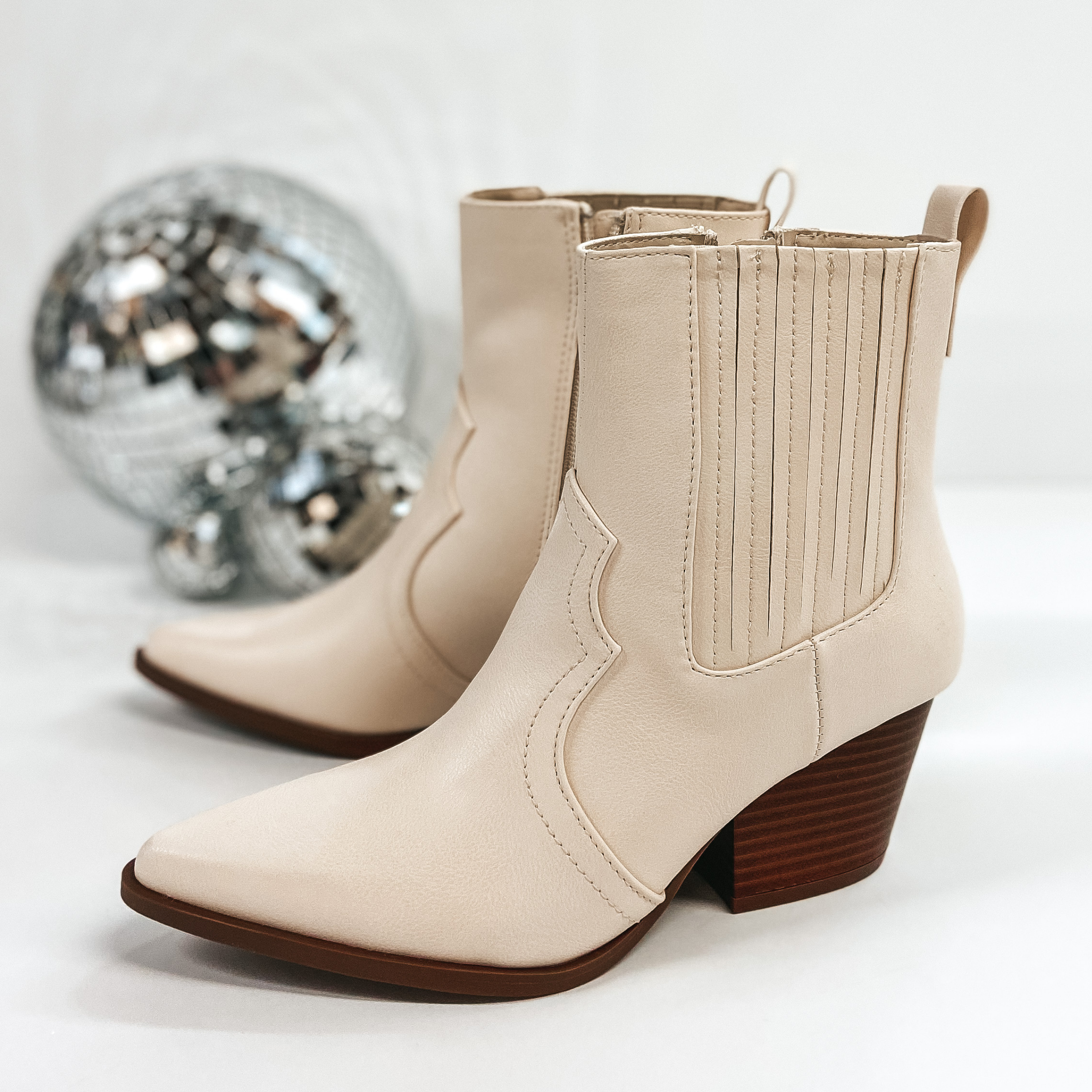 Western style short booties that are ivory. These shoes have a brown sole and heel. Pictured on white background with disco balls.