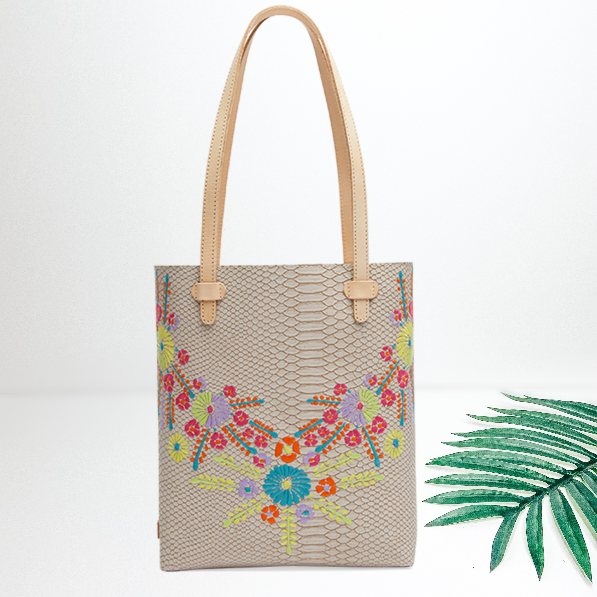 An ivory snakeskin tote bag with colorful floral embroidery. Pictured on white background with a green palm leaf.\