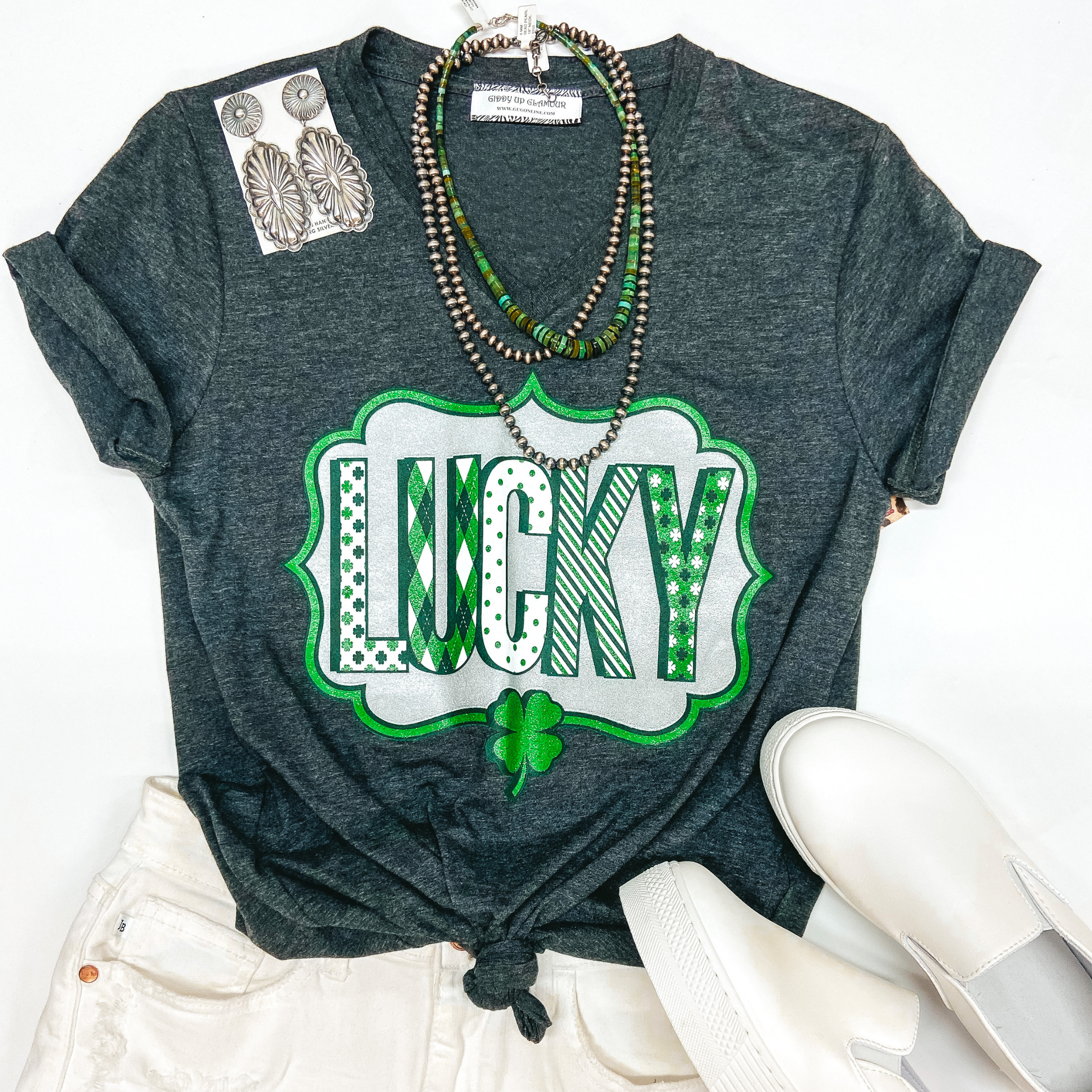 A grey v neck tee shirt with a graphic that says "Lucky". Pictured on white background with white sneakers, white shorts, and silver jewelry.