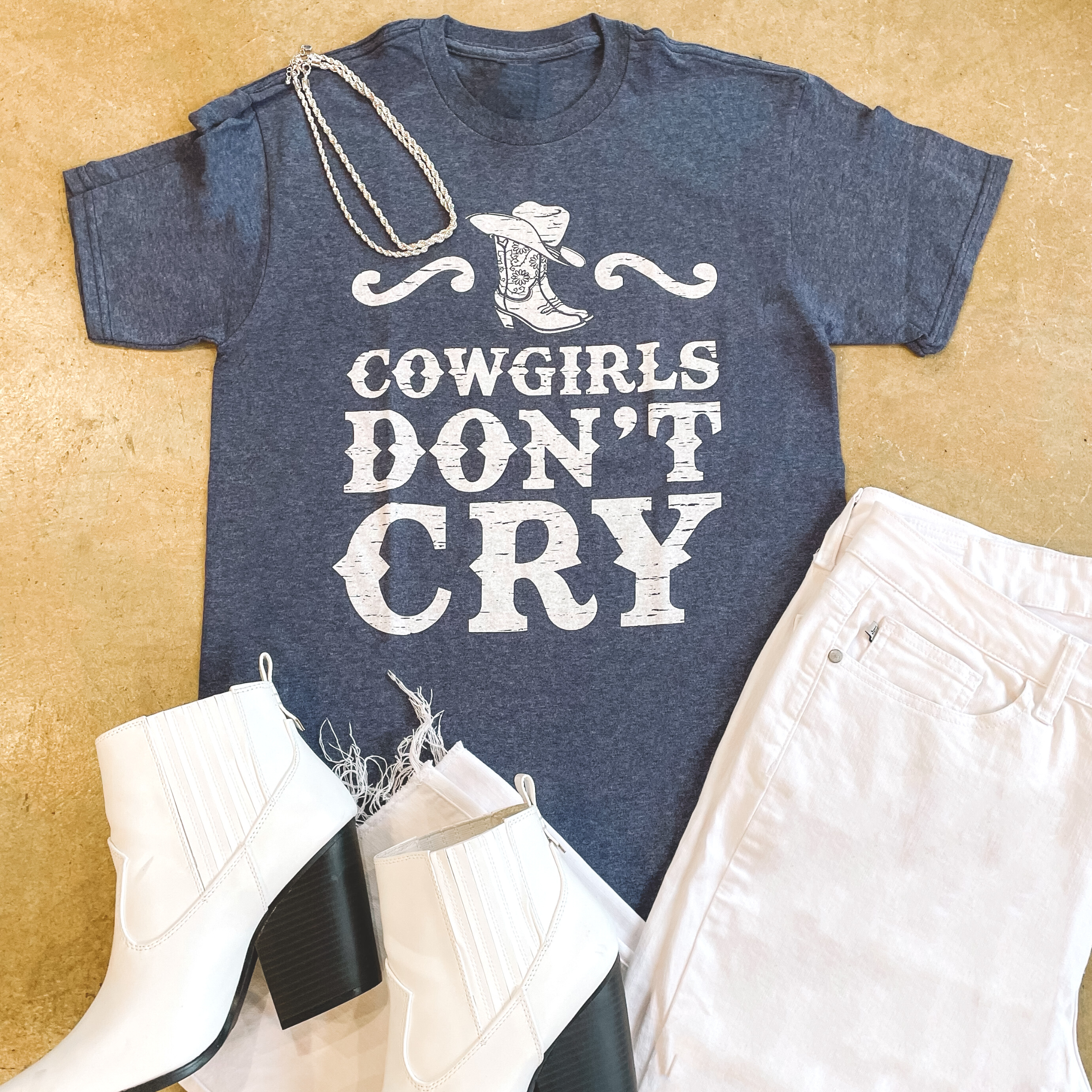 Denim color shirt with text saying "Cowgirls Don't Cry" and a boot decal on the top. Paired with the shirt in the top left is a double strand silver necklace and in the bottom left white booties along with white croped pants in the bottom right. Background is tan. 