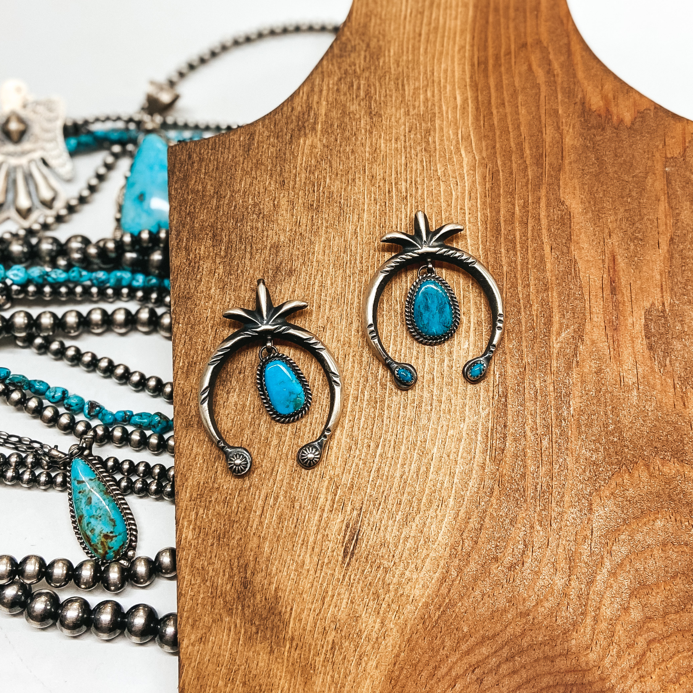 Two sterling silver Naja shaped necklace pendants with silver tooling and turquoise center dangles. Pendants are pictured on a wooden background with Navajo pearls and turquoise jewelry.