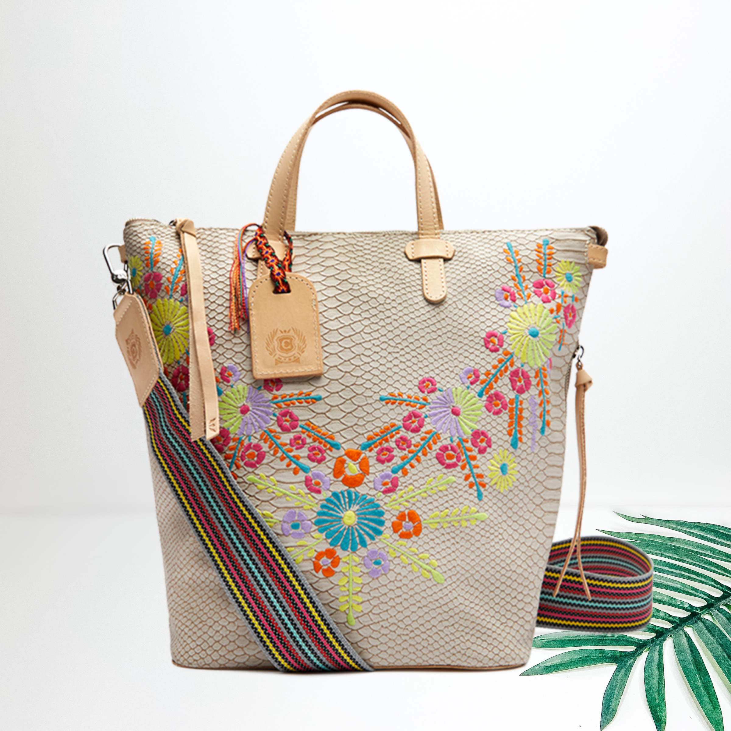 A beige snake print sling bag with colorful floral embroidery. Pictured on white background with a palm leaf.