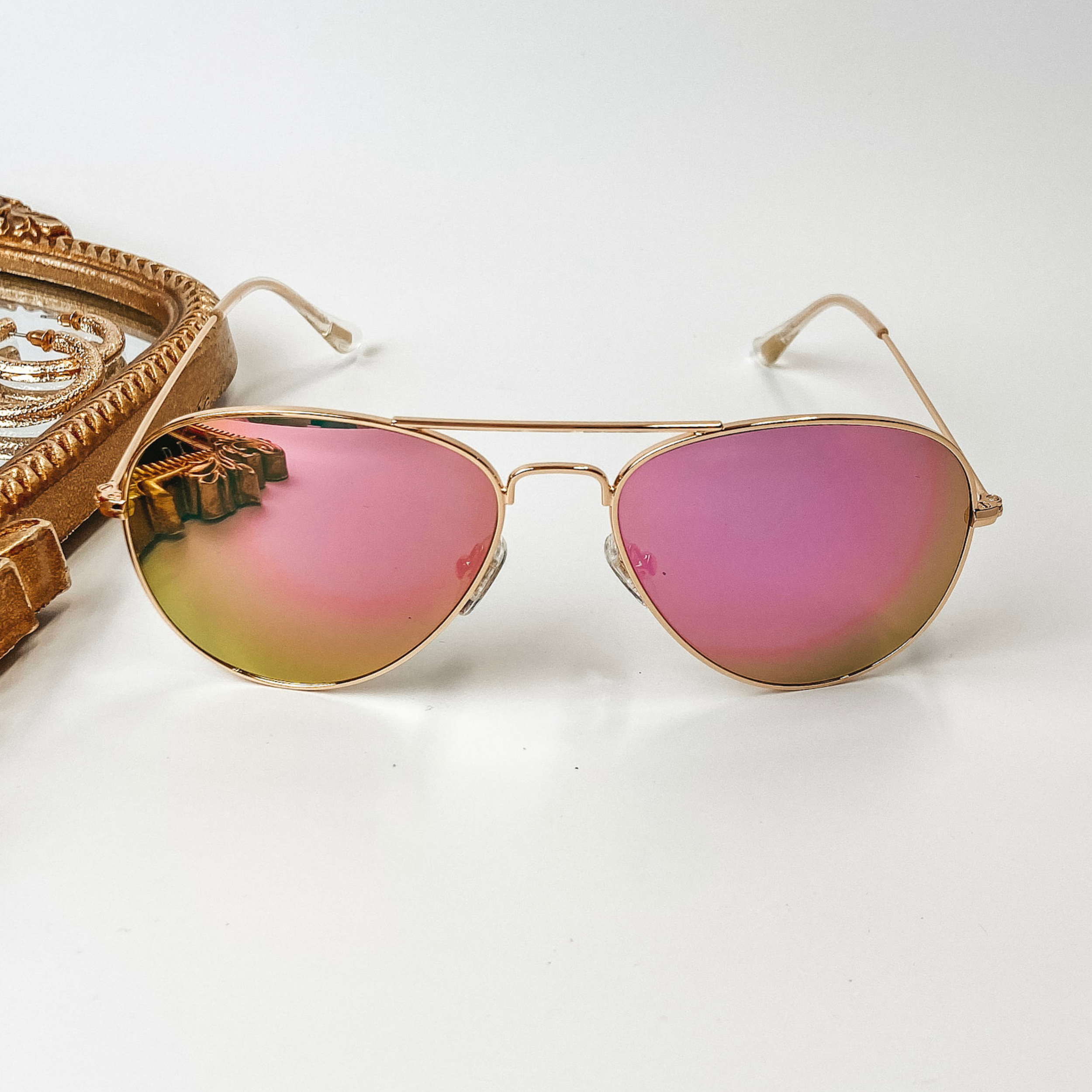  A pair of aviator style sunglasses with pink mirror lenses and gold tone frames. These sunglasses are pictured on a white background with gold jewelry.