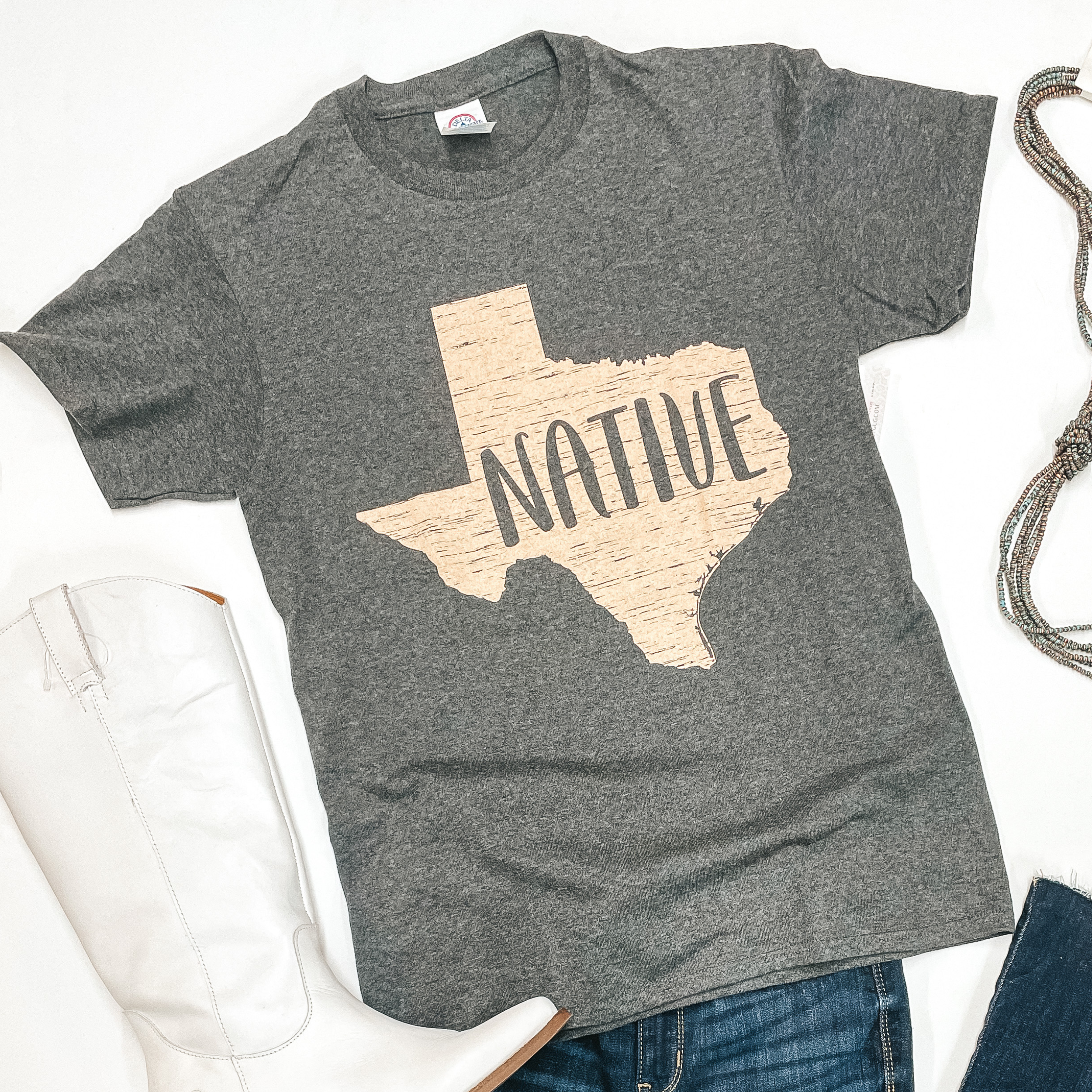 A gray tee is centered in the middle of the picture on a white background. In the center of the tee is a cream Texas with "native" in block letters. On the bottom left of the picture is white boots and bottom right is jeans. Towards the right of the picture is a long strand of faux navajo necklaces. 