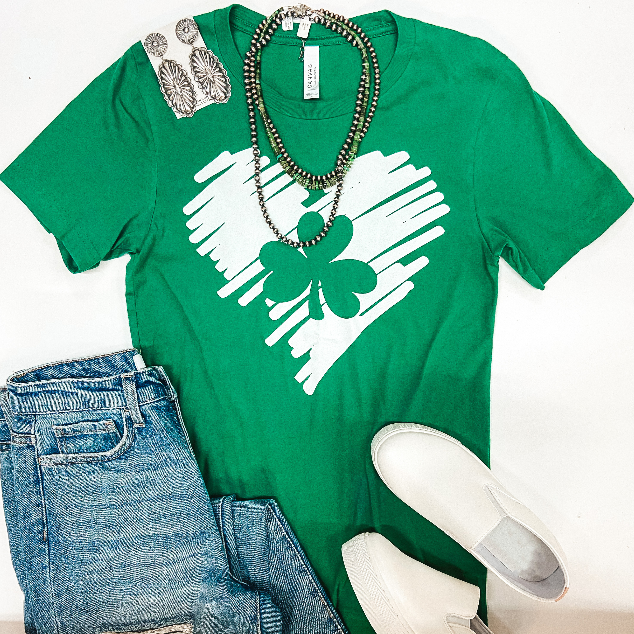 A green tee shirt with a white sketched heart and a clover outline. Pictured with white sneakers, silver jewelry, and light wash distressed jeans.