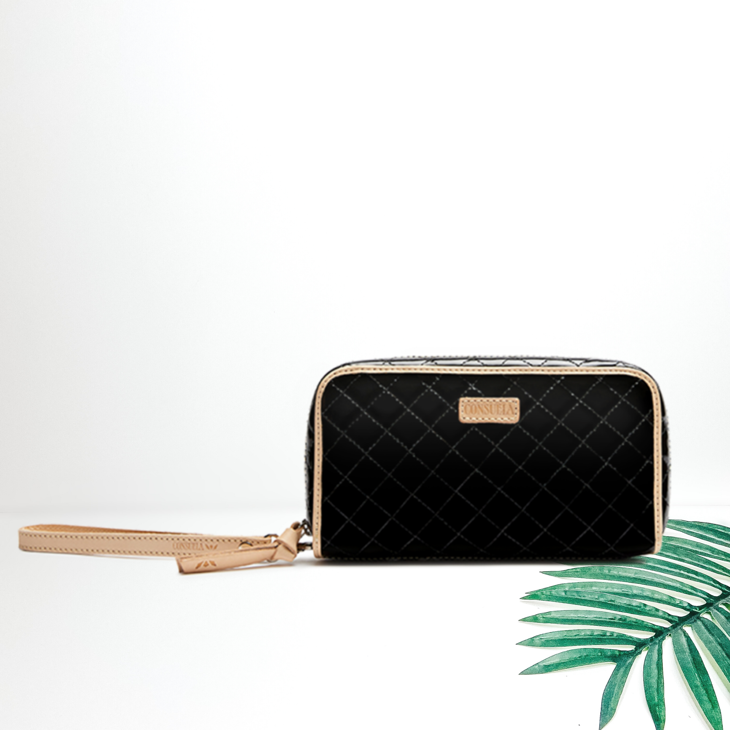 A black quilted wallet with leather trim pictured on a white background with a palm leaf.