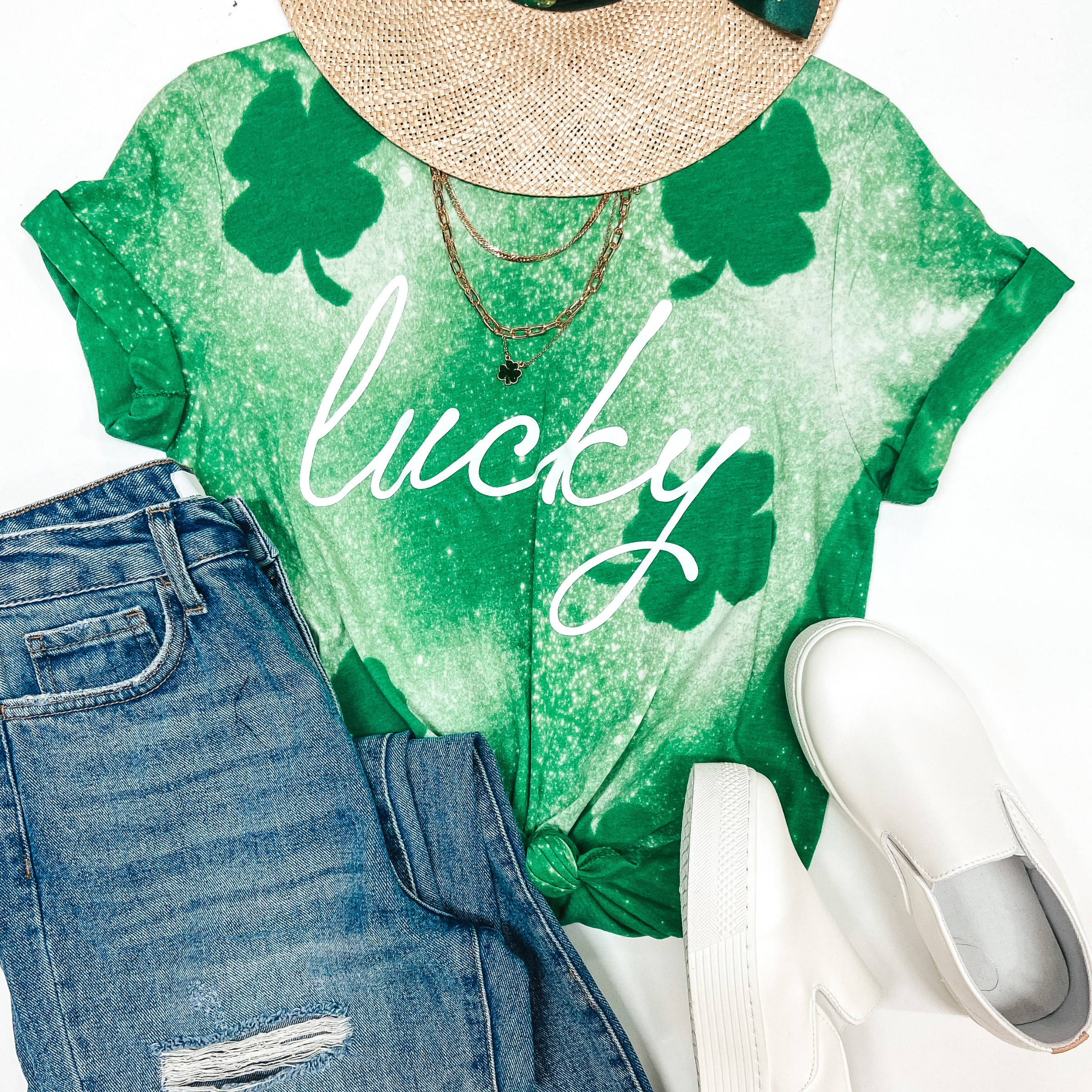 A green tee shirt with bleach splatter distressing and clovers that says "Lucky." pictured on white background with light wash jeans, white sneakers, and gold jewelry.