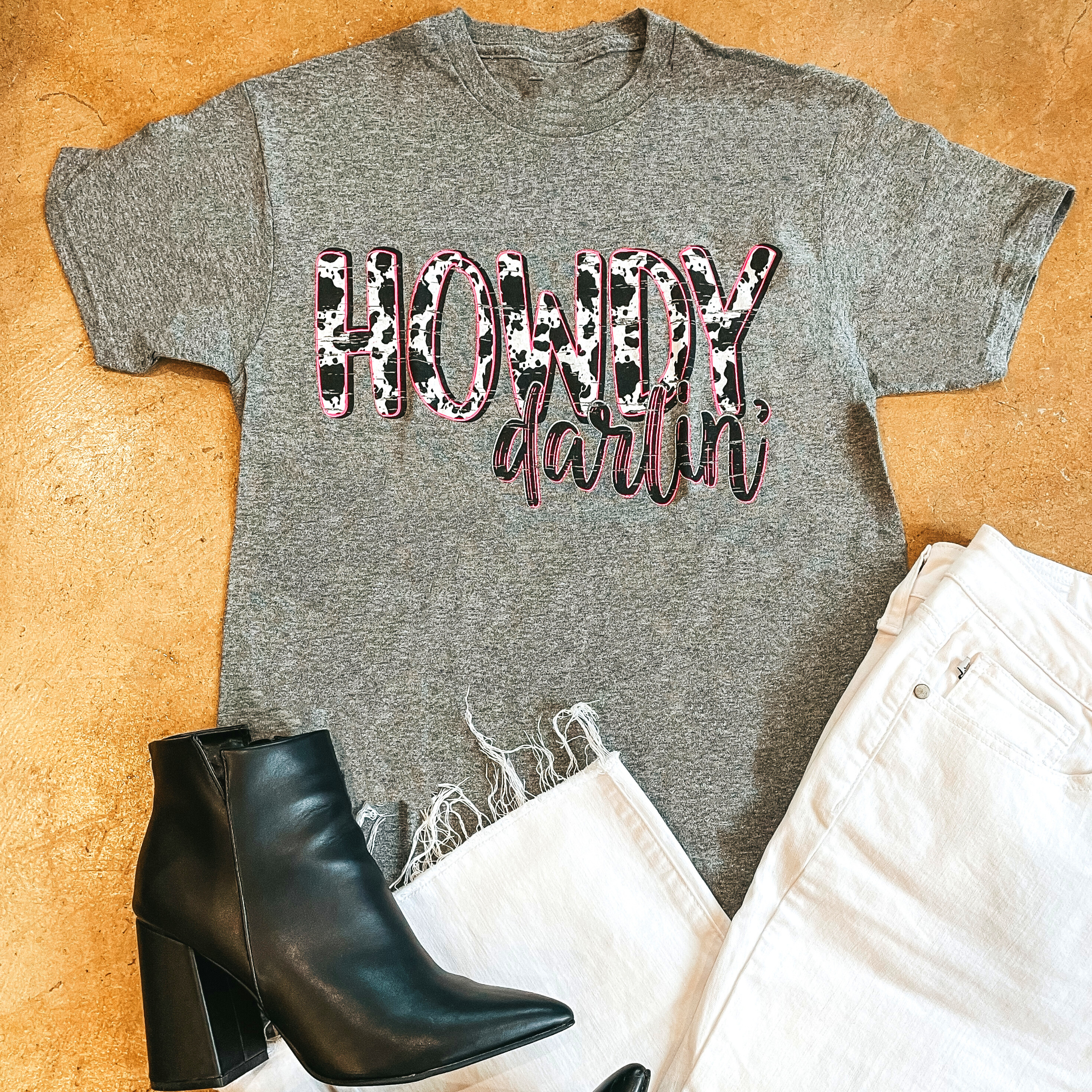 "Howdy Darlin" written on a gray short sleeve t-shirt with "Howdy" in cowhide and all outlined in pink. Black booties are towards the bottom left of the screen and white bants are on the bottom right. Background is brown. 