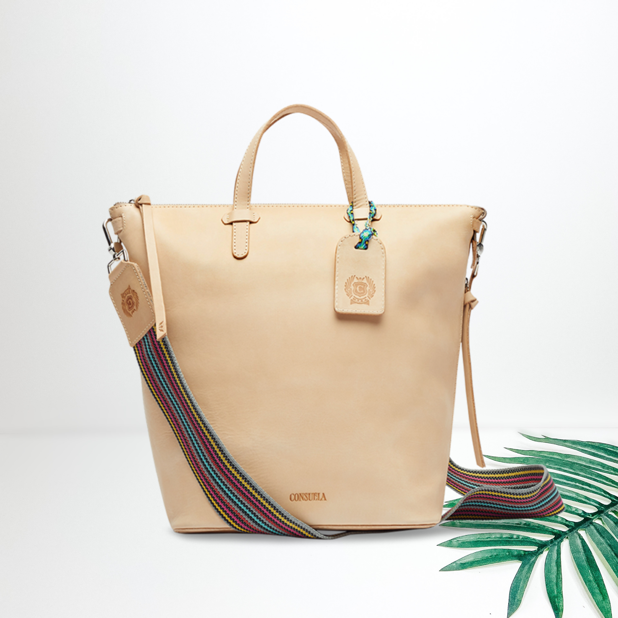 A light tan leather bag with a white a crossbody strap. Pictured on a white background with a palm leaf.
