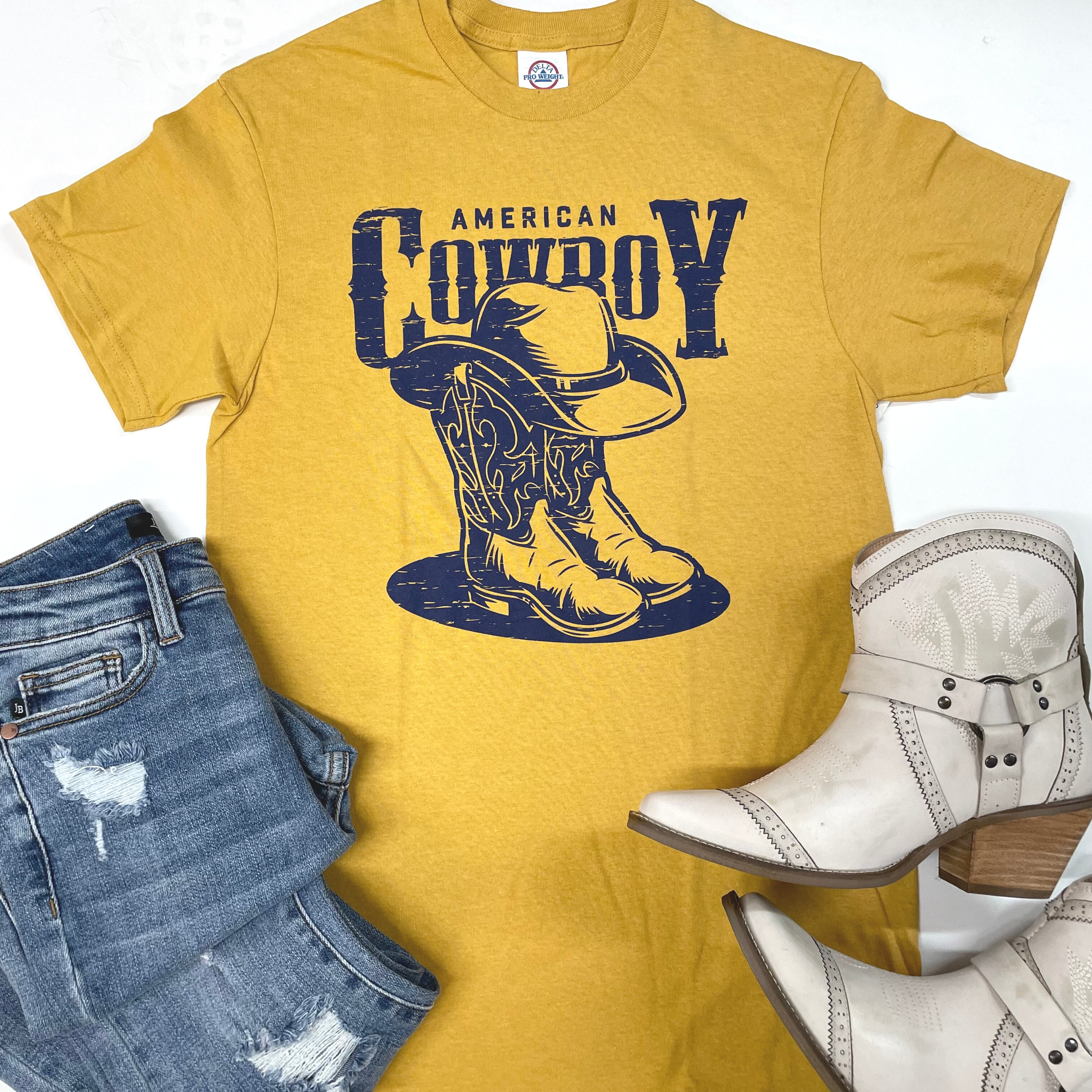 A mustard yellow tee is in the center of the picture on a solid white background. The tee has "American Cowboy" written in the center, with cowboy boots and a hat in navy. On the bottom left of the picture is a light wash pair of jeans and on the bottom right is a white pair of jeans. 