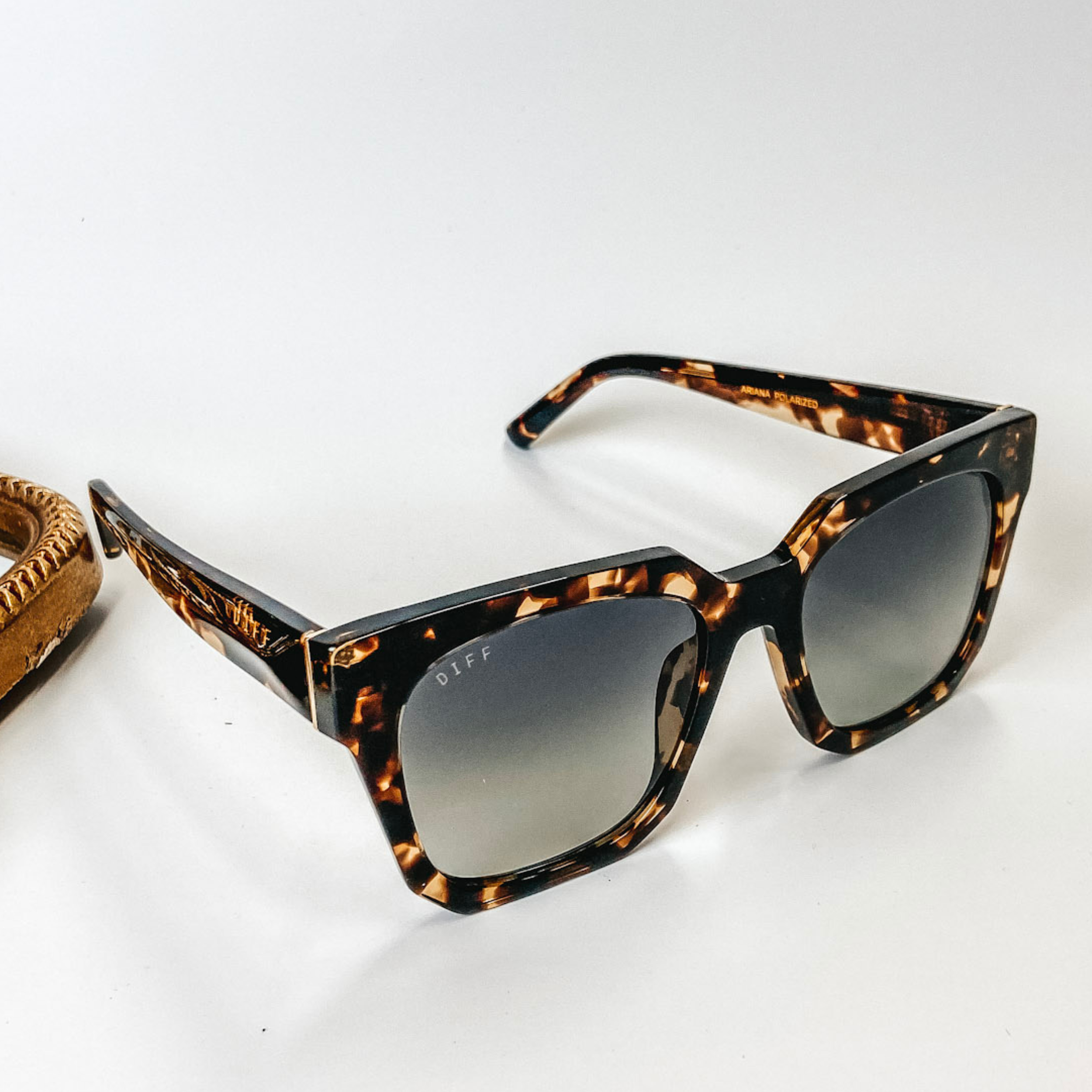 A pair of tortoise print frame sunglasses with gradient square lenses in black. These sunglasses are pictured on a white background with gold jewelry.