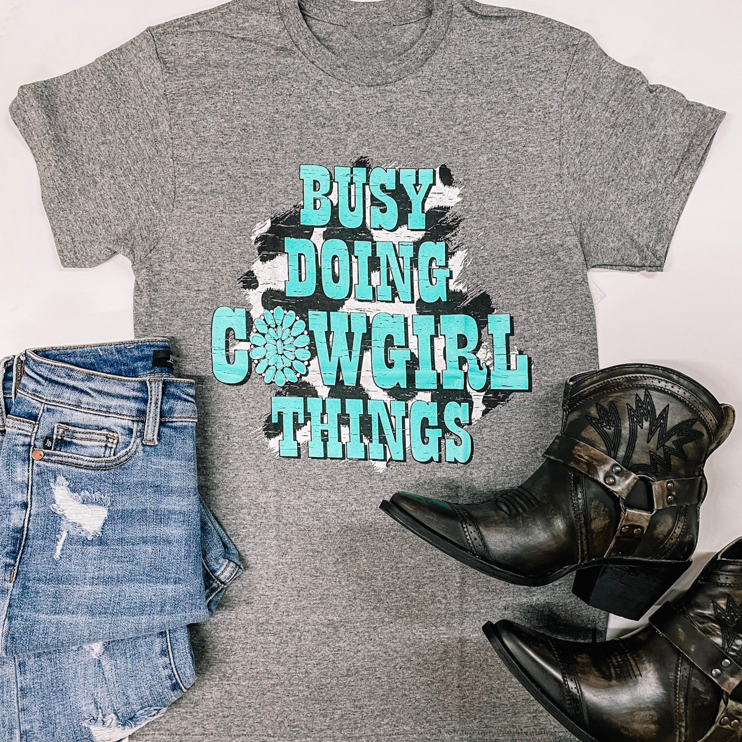 A grey tee is laid out on a white background. In the middle of the tee is a cowprint circle under turquoise letters that state, "Busy doing cowgirl things." Jeans are laid out on the bottom left corner and on the bottom right is a black pair of boots.  