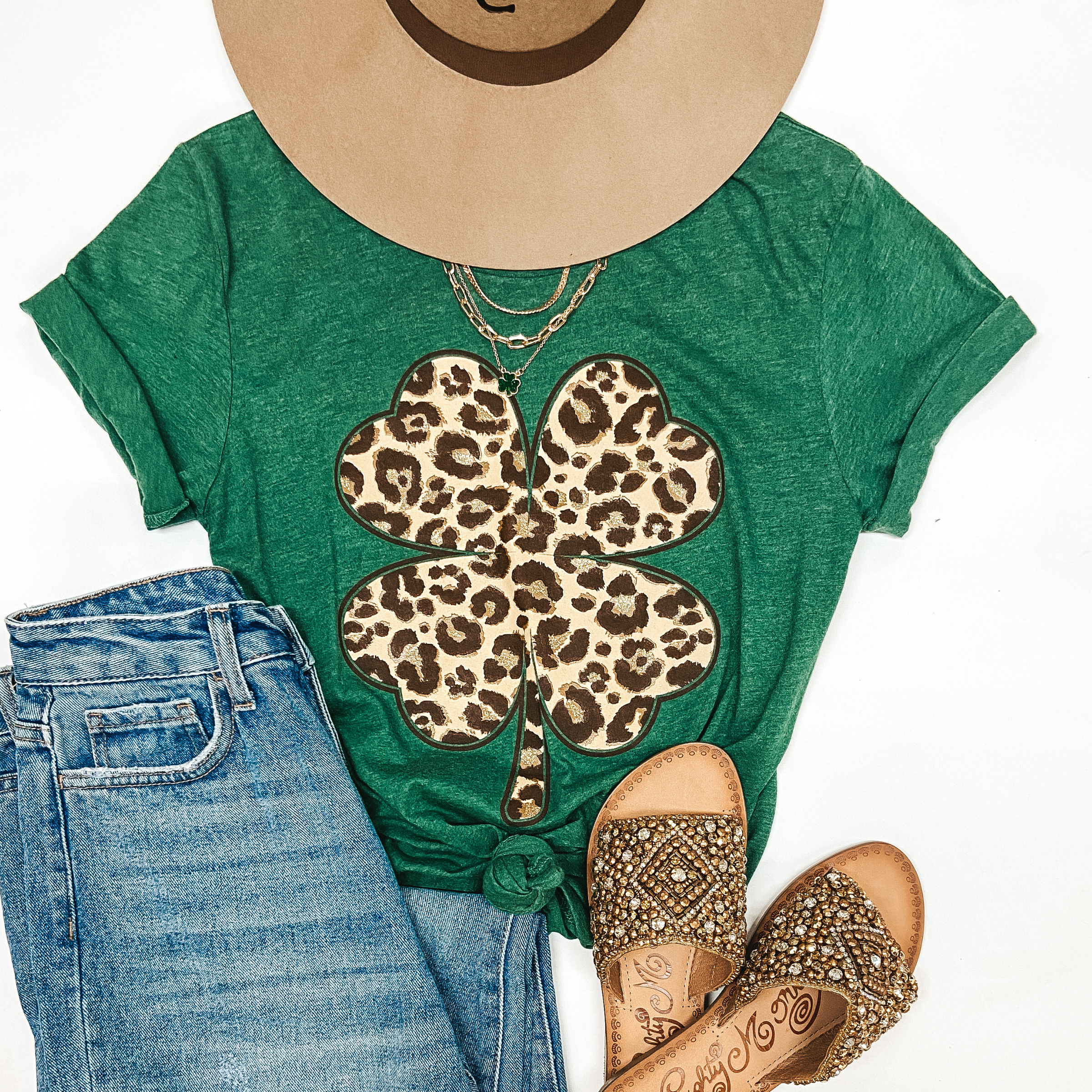 A green tee shirt with a leopard print clover. Pictured with light wash jeans, a tan hat, and gold sandals.