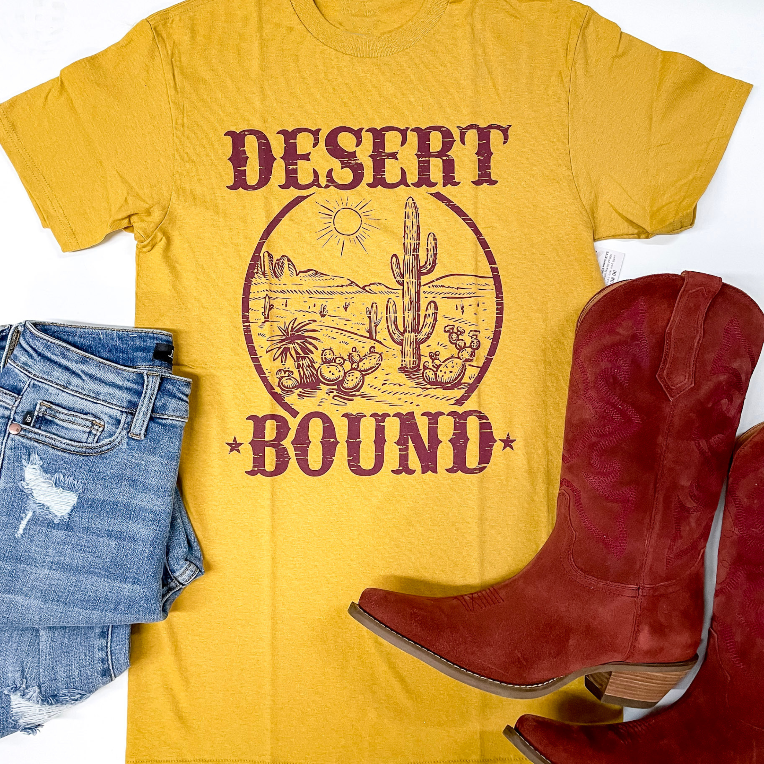 A mustard yellow tee is laid center on a white background. In the center of the tee is a graphic design of a desert with the words, "desert bound." On the bottom left corner is a light-wash pair of jeans and on the bottom right there is a red pair of boots. 