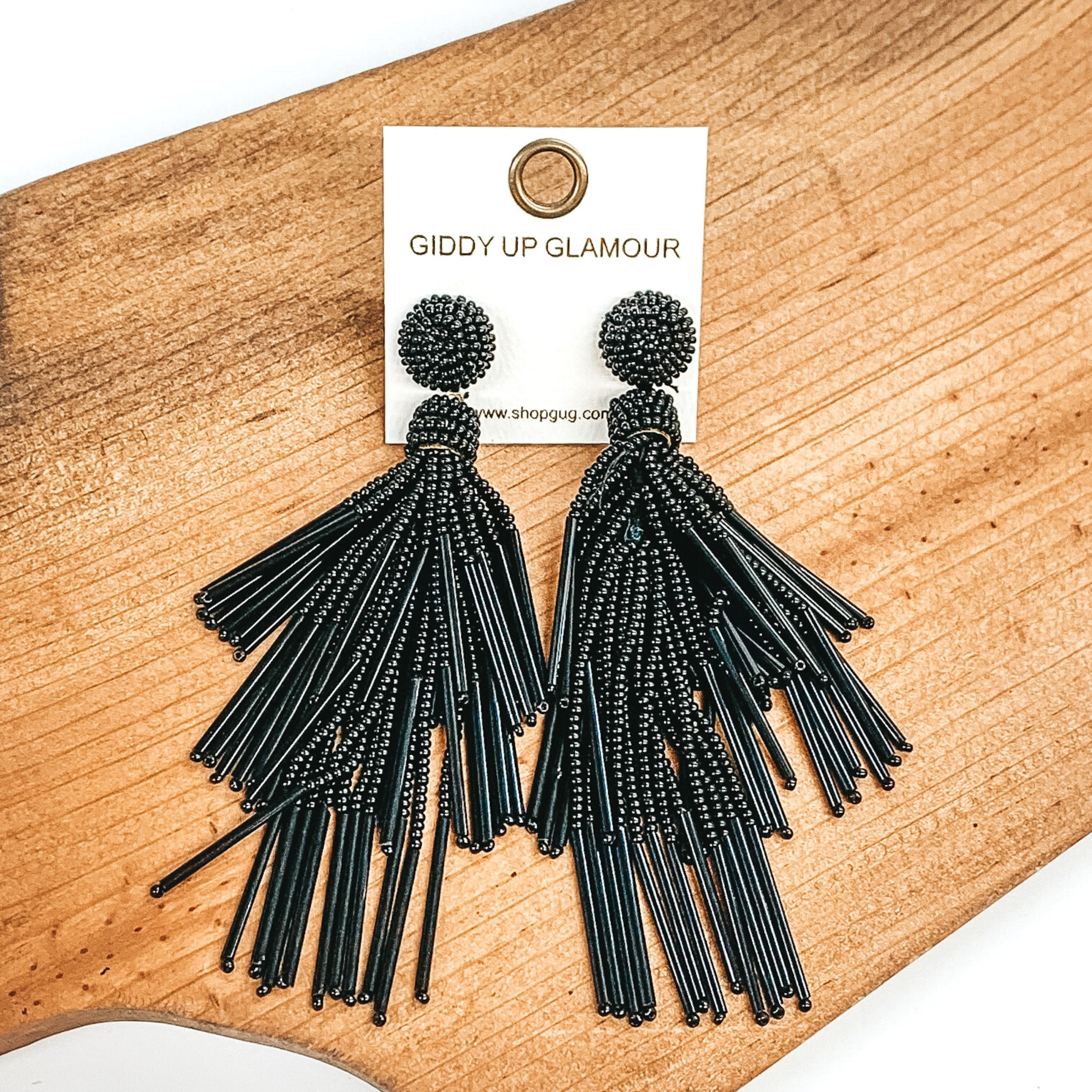 Circle beaded stud earrings with a beaded tassel in black. These earrings are pictured laying on a brown piece of wood on a white background. 