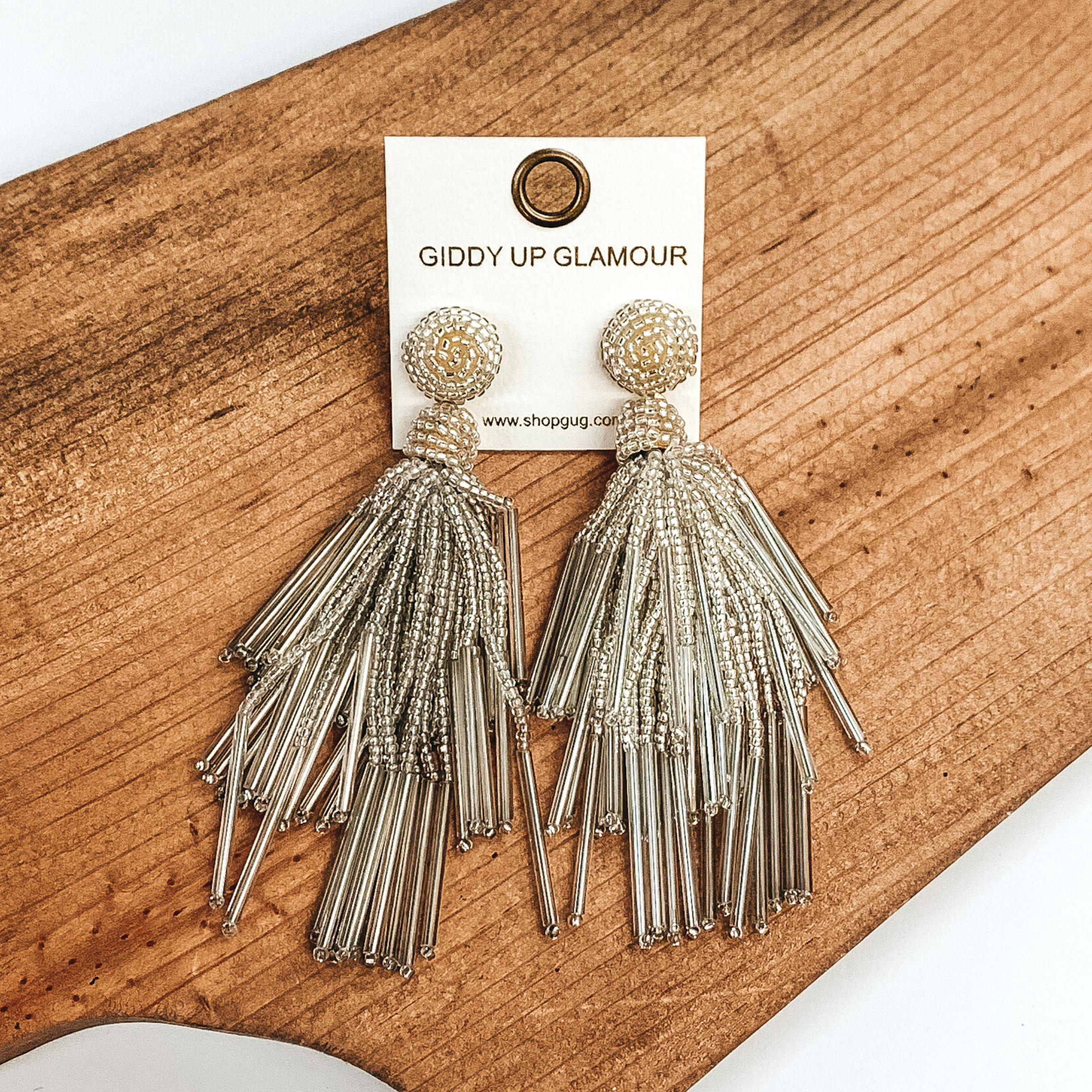 Circle beaded stud earrings with a beaded tassel in silver. These earrings are pictured laying on a brown piece of wood on a white background.