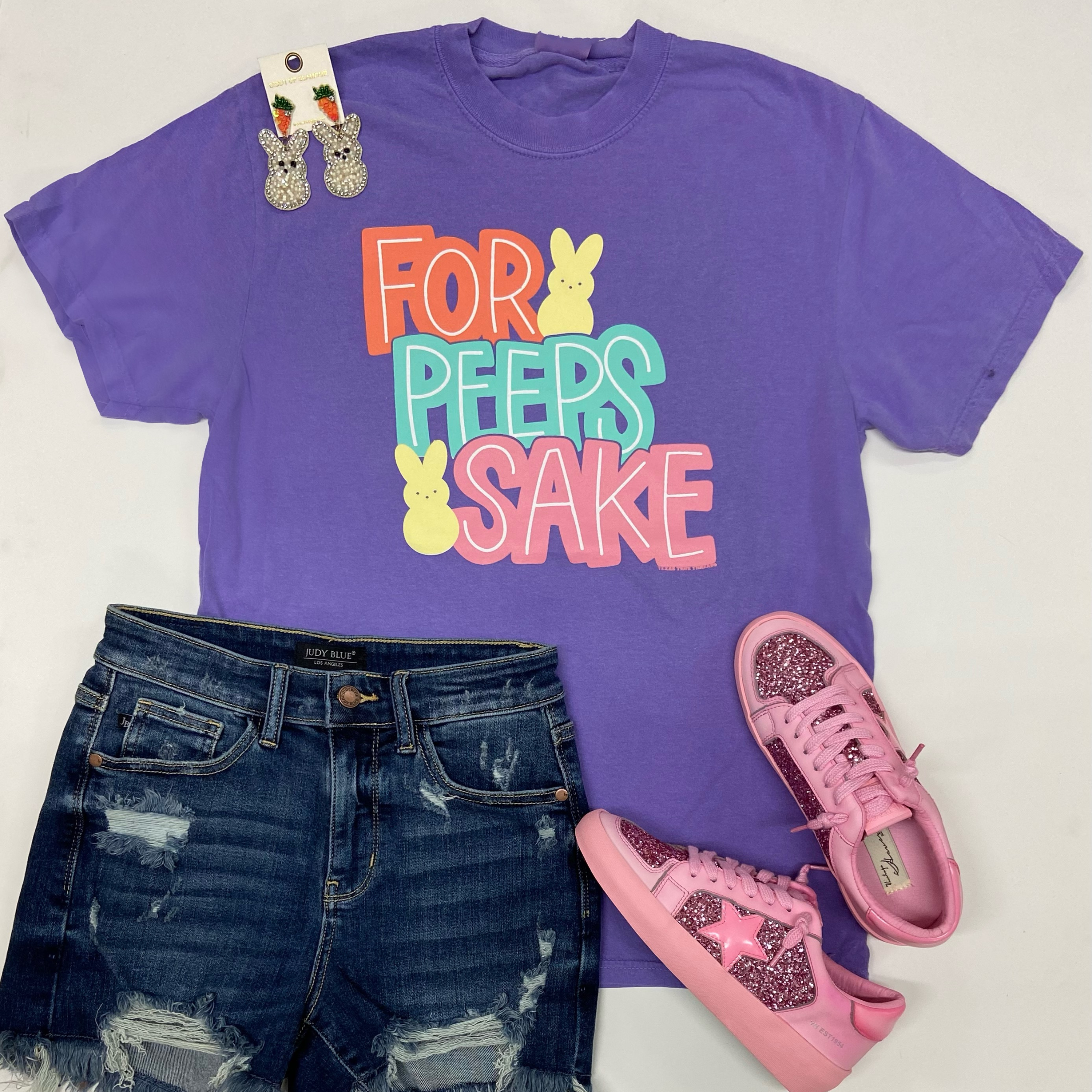 A purple graphic tee shirt that says "For Peeps Sake" with yellow peeps. Pictured on a white background with pink sneakers, distressed shorts, and peep earrings.