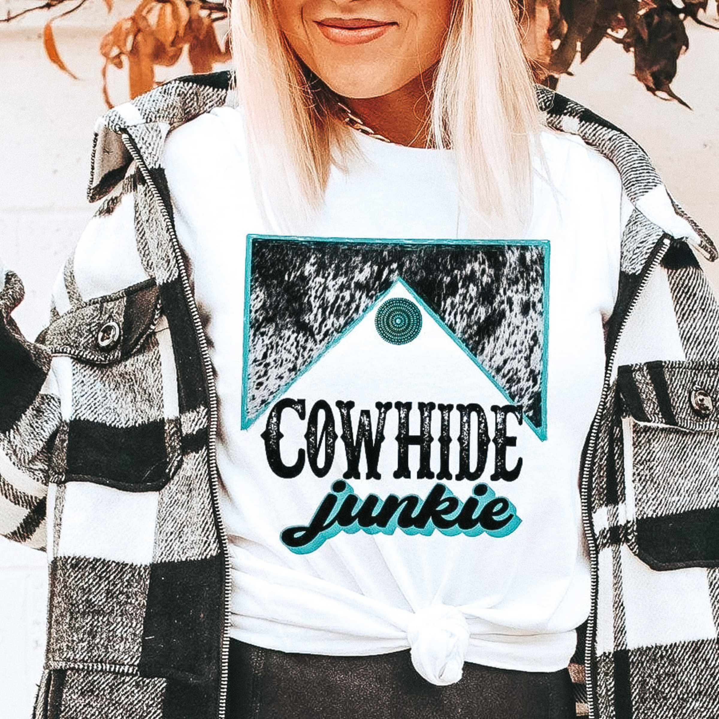 Model is wearing a white graphic tee that says "Cowhide Junkie" with a turquoise and cowhide graphic. Model has it paired with a black and white buffalo plaid jacket.