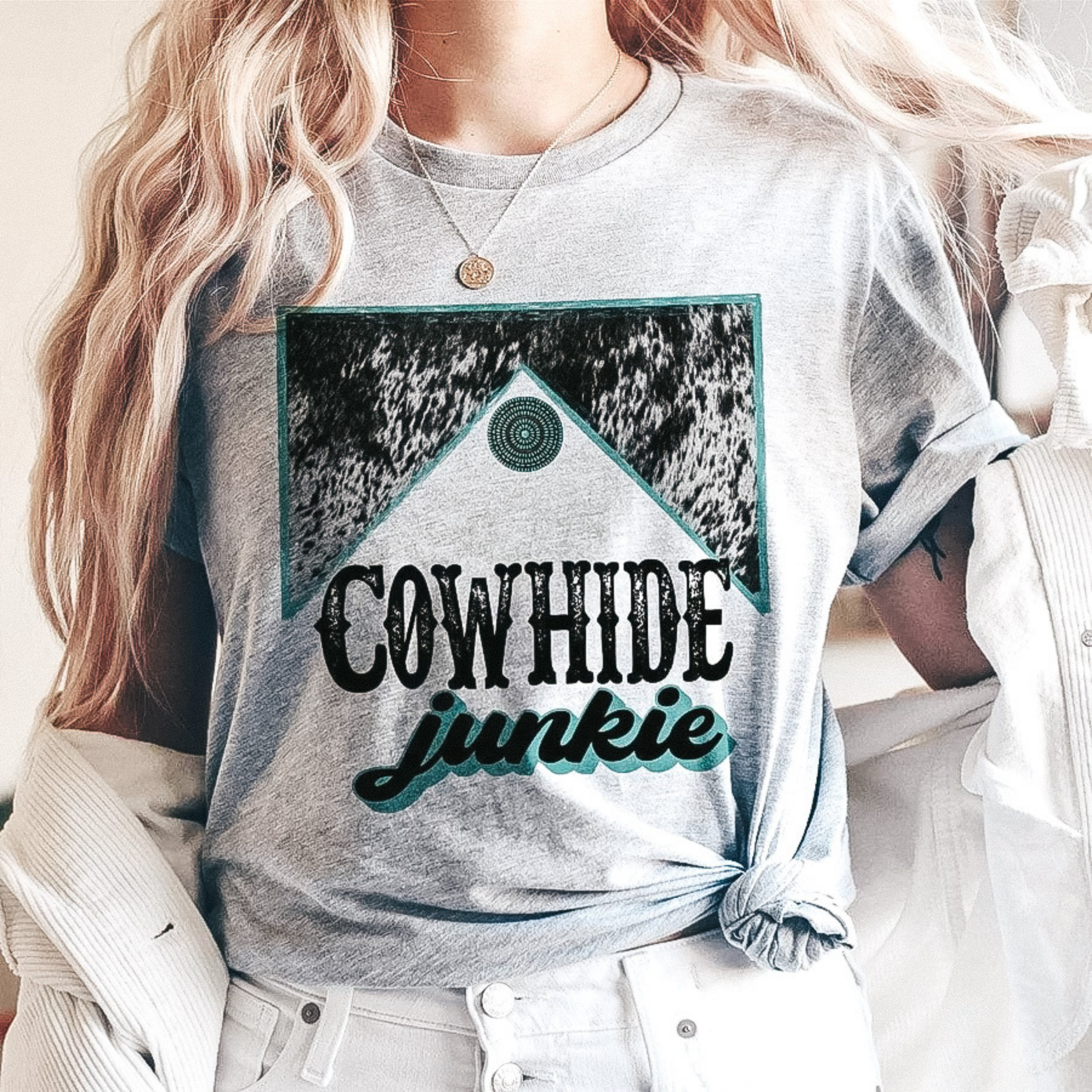 Model is wearing a grey tee shirt that says "Cowhide Junkie" with a turquoise and cowhide graphic. Model has it paired with a white corduroy jacket and white pants.