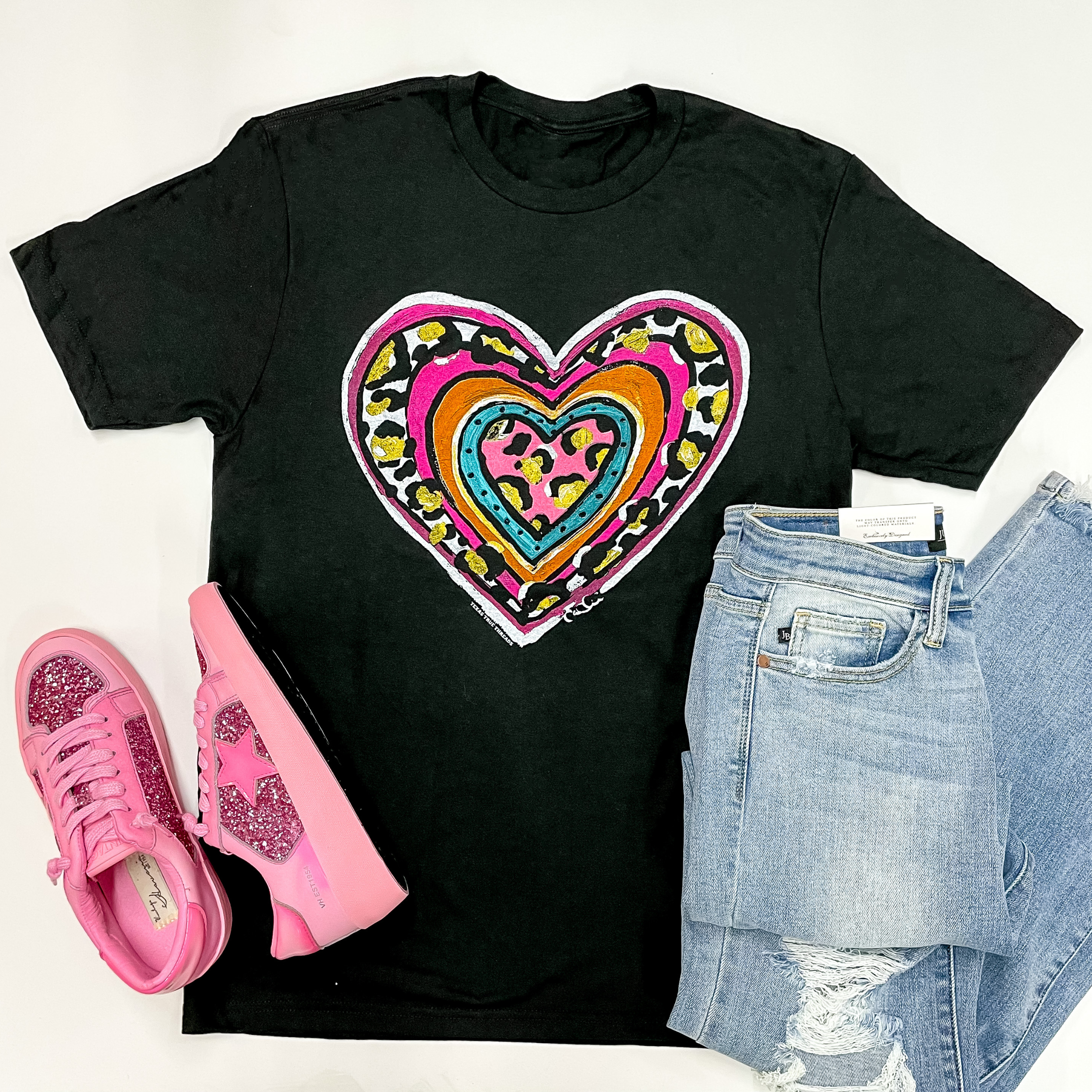 A black short sleeve tee shirt with a heart graphic that is  a mix of blue, pink, orange, gold, and leopard print. This tee shirt is pictured on a white background with distressed jeans and pink sneakers to match.