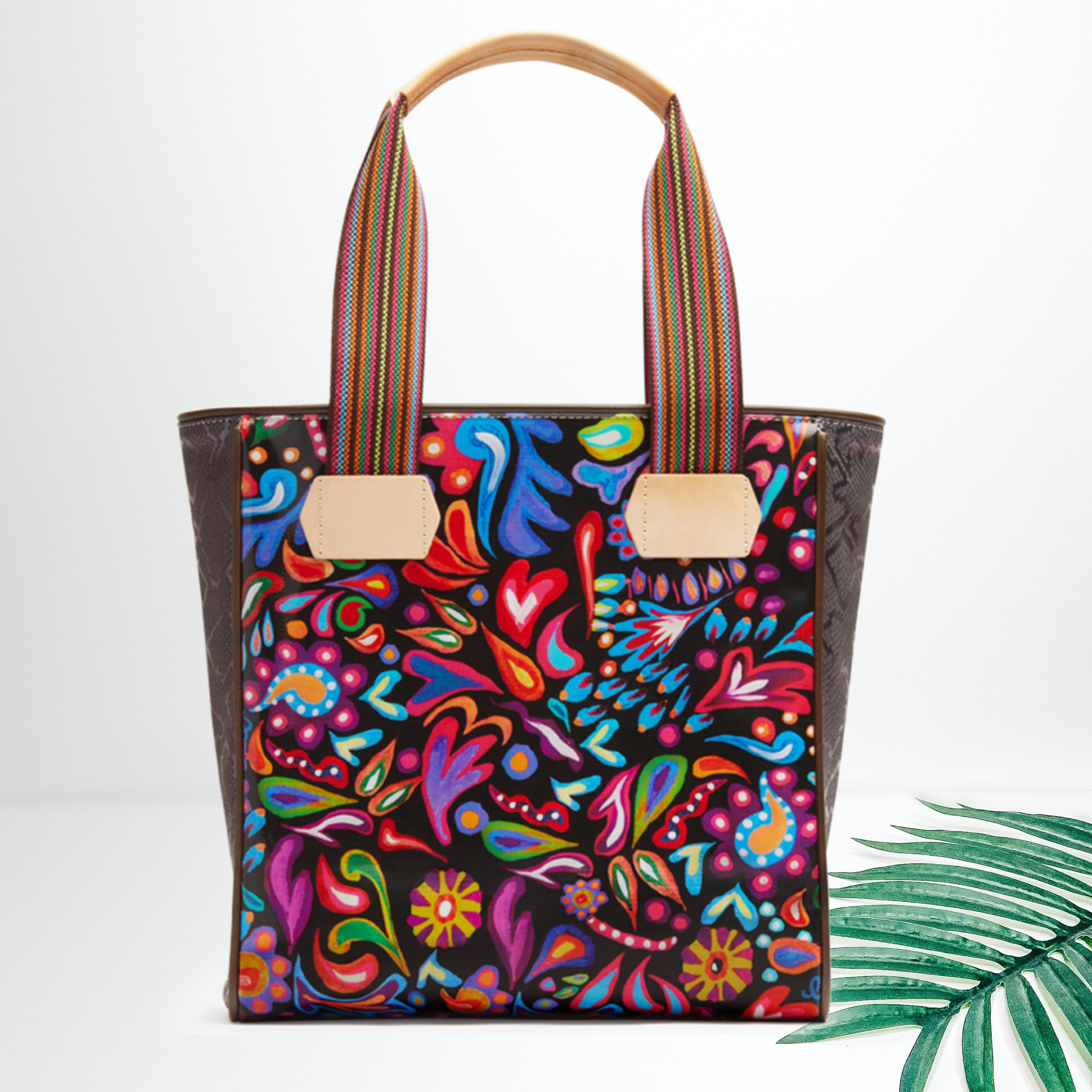 A large shoulder tote that is black with a colorful paisley print. Pictured on white background with a palm leaf.