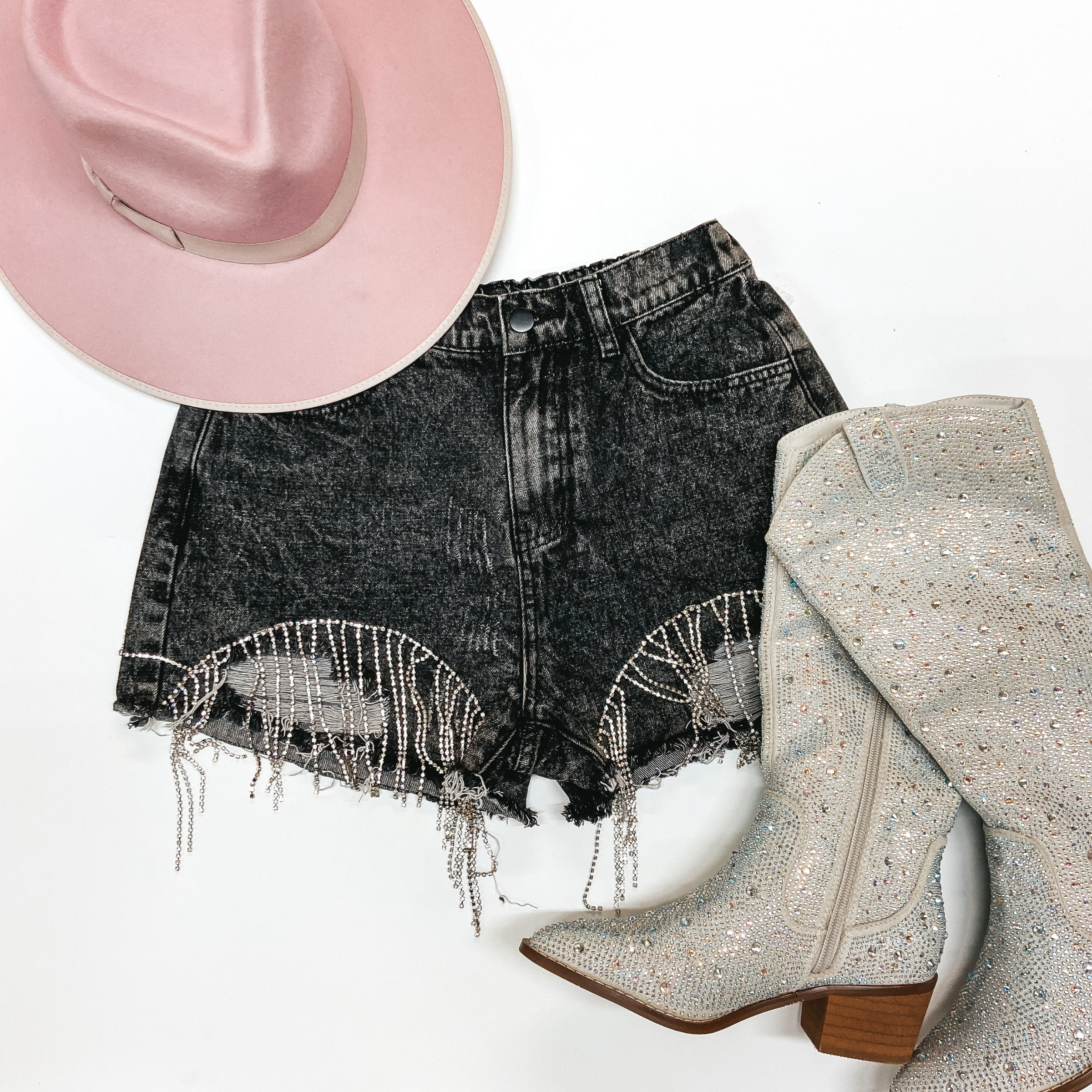 Black shorts with crystal fringe on the bottom on a white background. Paired with a pink Charlie 1 Horse hat in the top left and Silver rhinestone booties on the bottom right. 