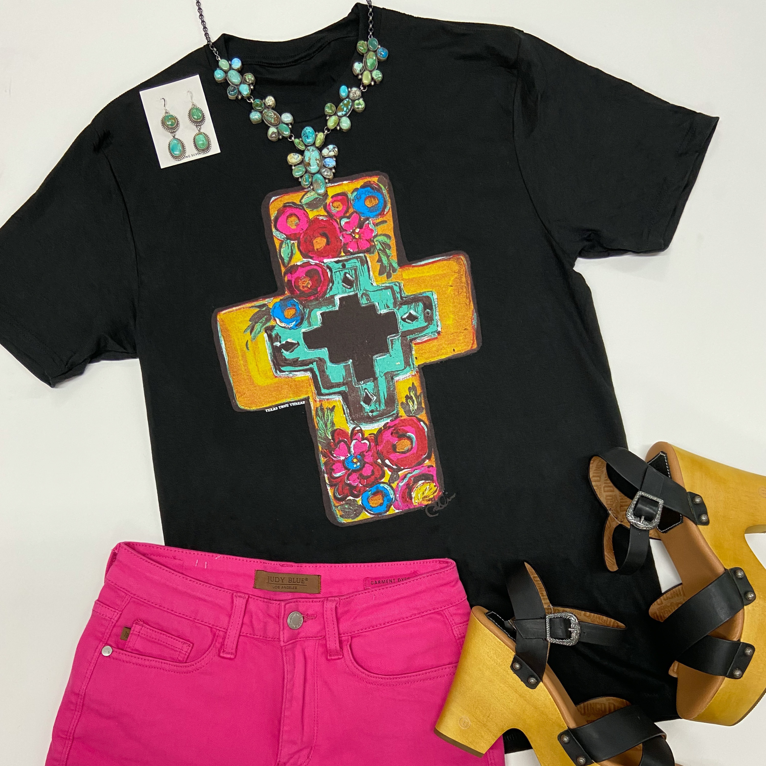 A black tee shirt with a crew neckline, short sleeves, and a graphic of a mustard yellow cross with flowers. This tee is pictured on a white background with pink shorts, black heels, and Navajo jewelry.