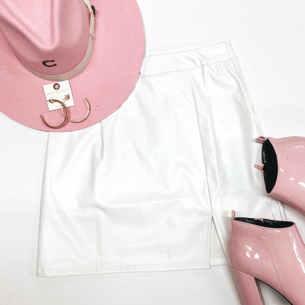 A white leather skirt that has a side slit. Pictured with a pink hat, pink booties, and gold jewelry on a white background.