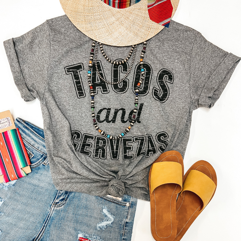 A heather grey graphic tee with the phrase "Tacos and Cervezas" in black. Pictured on white background with yellow sandals, denim shorts, sterling silver jewelry, a serape koozie, and a straw hat.