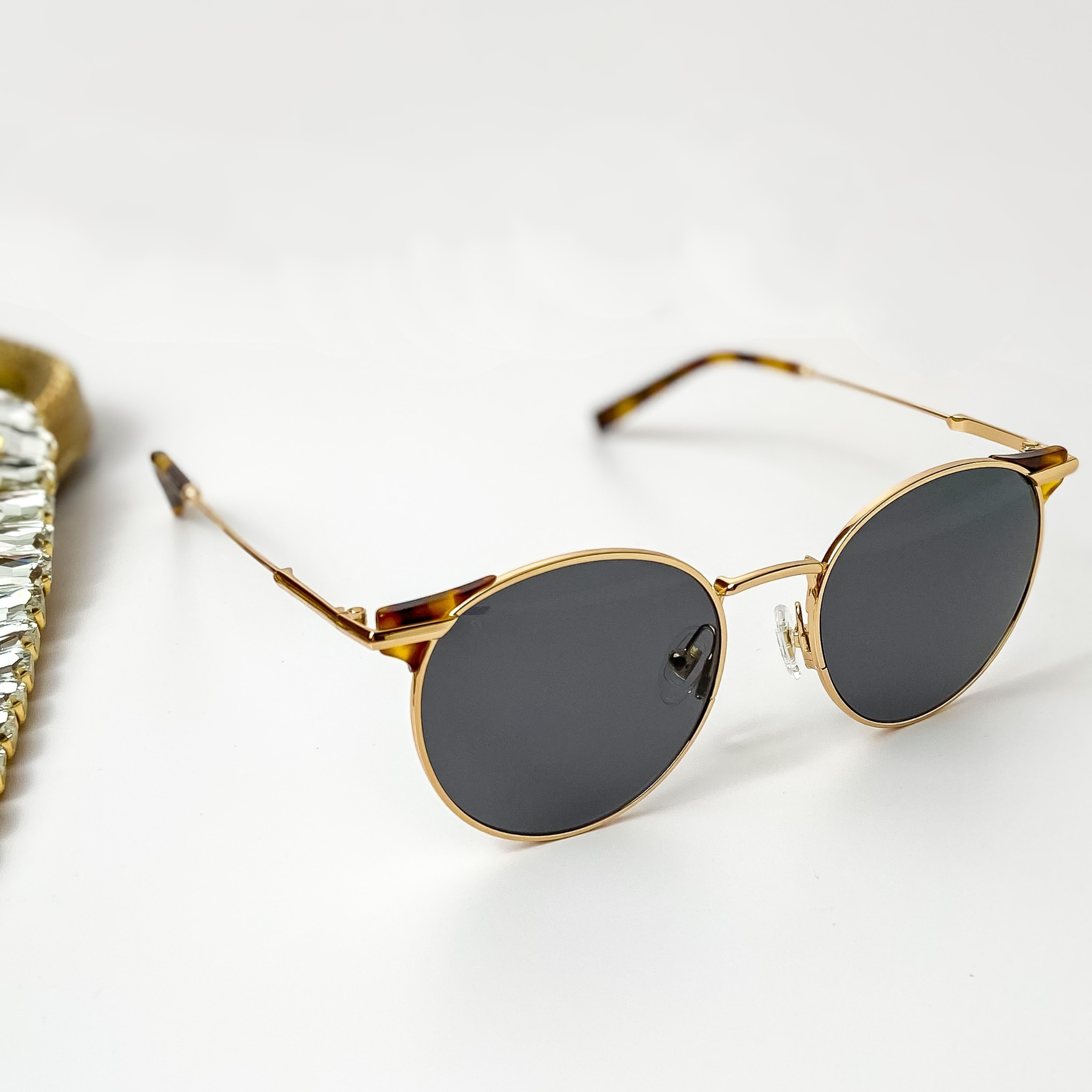 A pair of round sunglasses with grey lenses and gold tone frames. Pictured on a white background with gold jewelry.