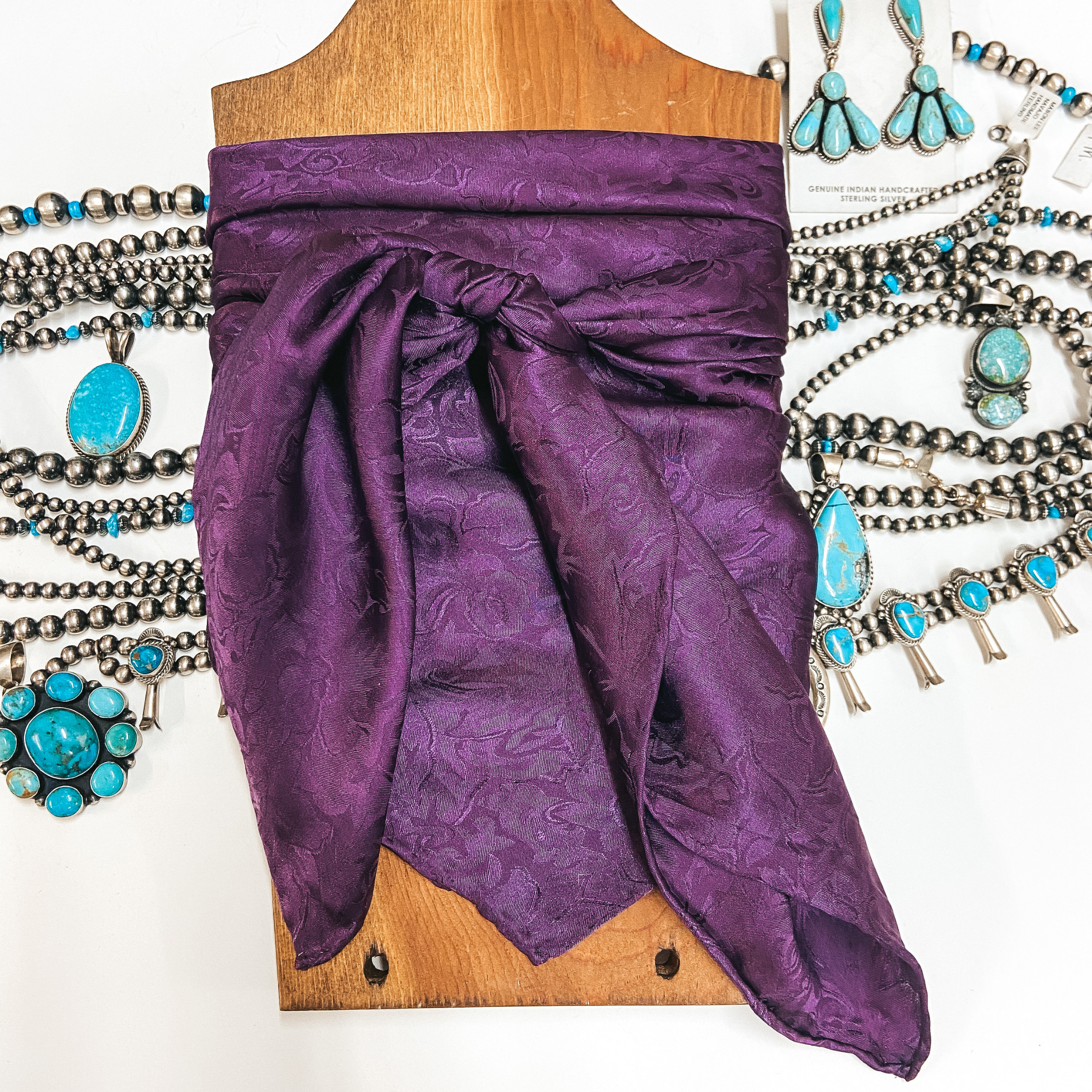 A jacquard print wild rag tied around a wooden display. Pictured on white background with sterling silver and turquoise jewelry.
