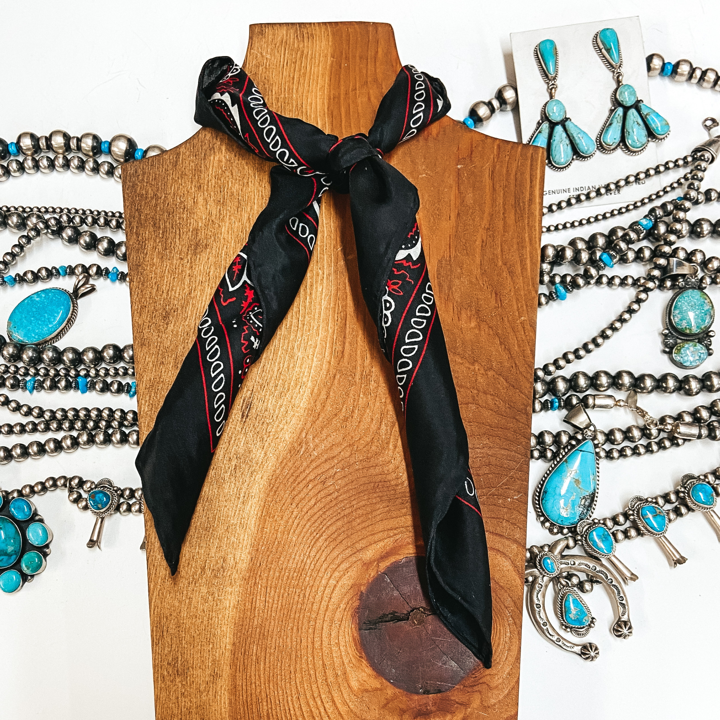 A black silky wild rag with red and white bandana print tied around a wooden display. Pictured on white background with silver and turquoise jewelry.