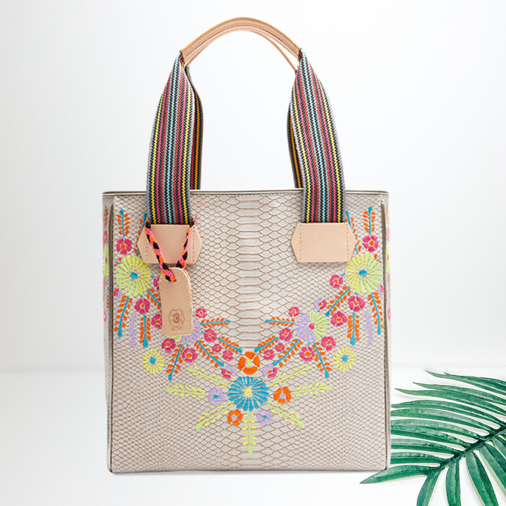 Centered in the picture is a cream colored tote with multi colored embroidery along the sides. To the right of the tote is a palm leaf, all on a white background. 