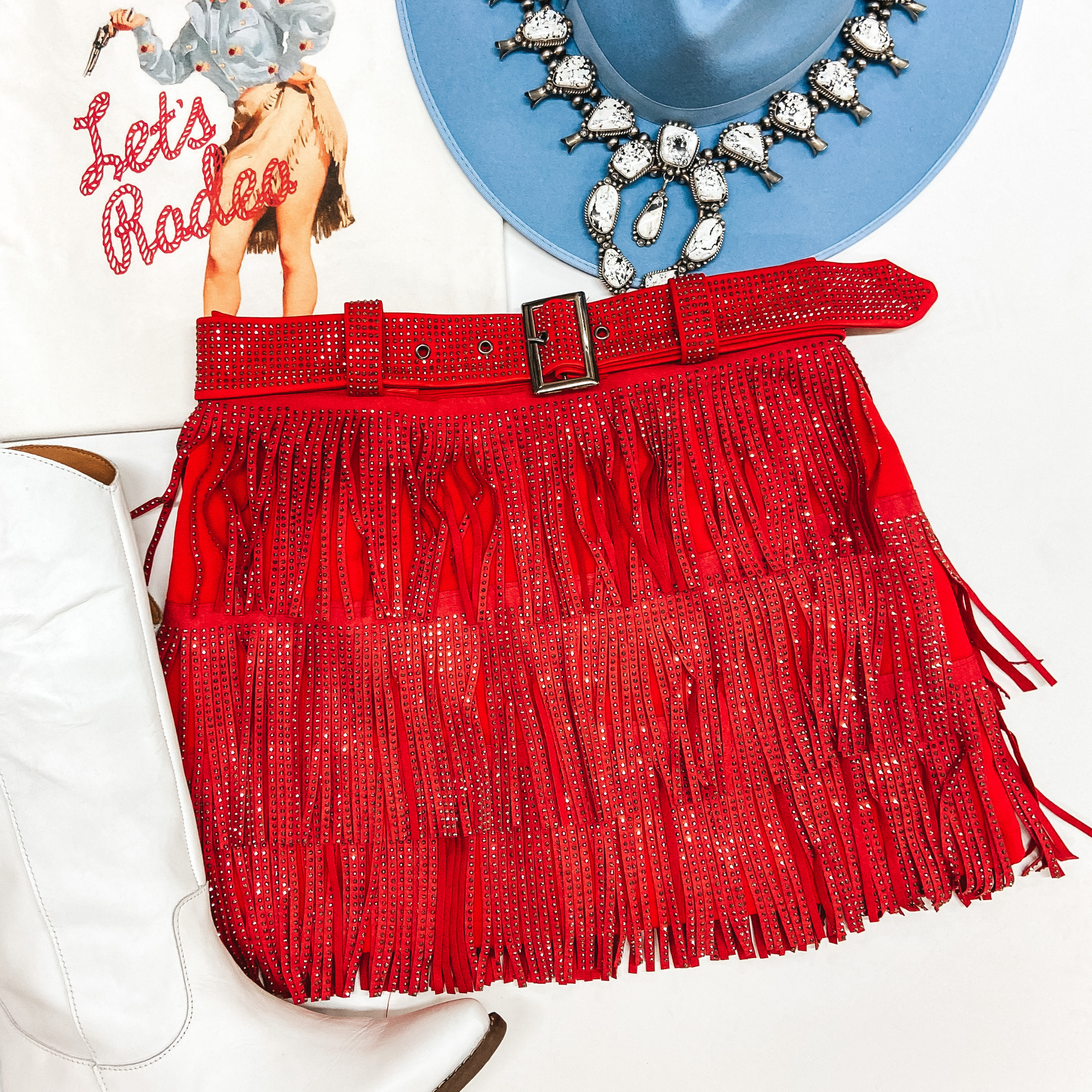A red fringe crystal skirt with a red crystal belt. Pictured with a white tee shirt, white boots, a blue hat, and white buffalo squash blossom.