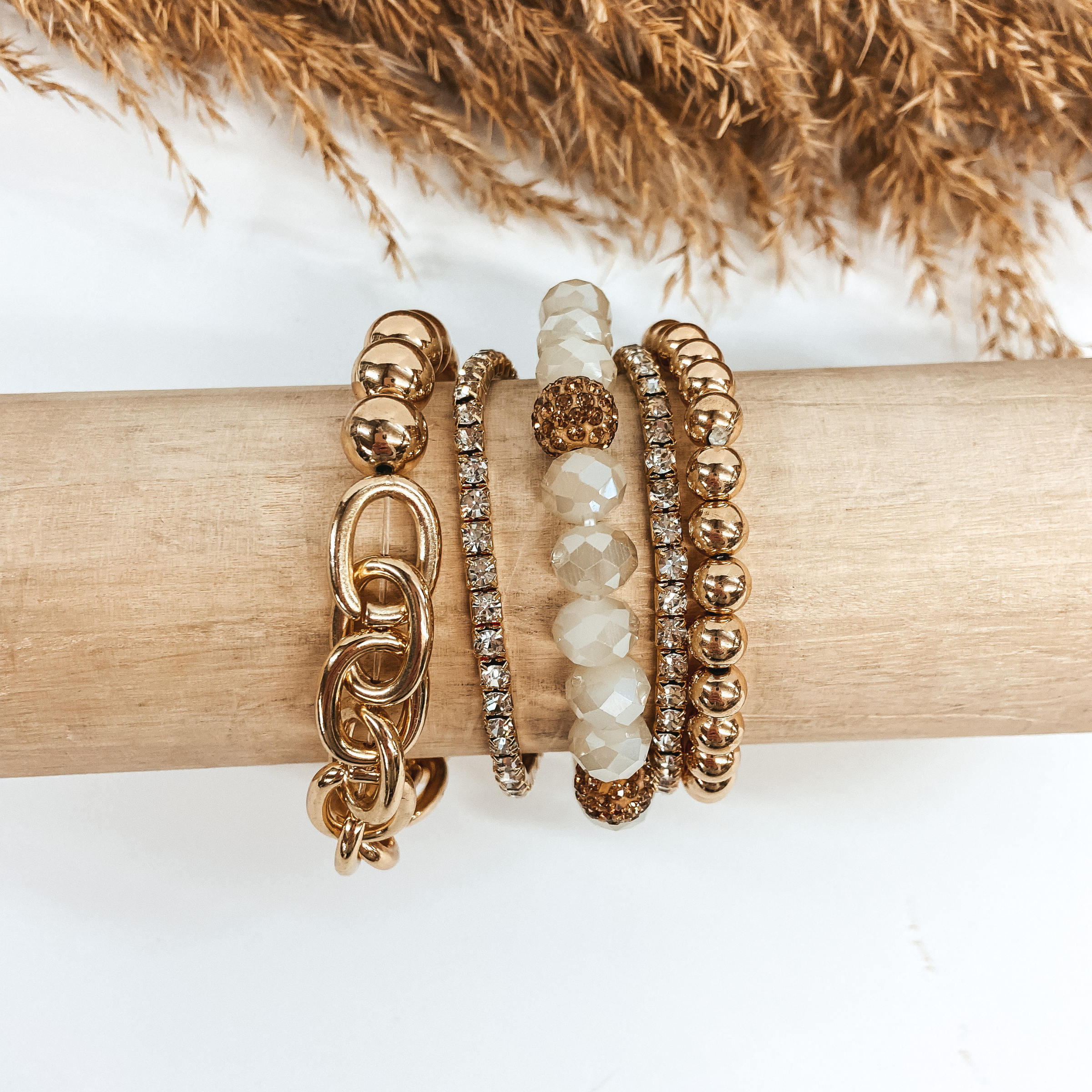  A mix of crystal and gold beaded bracelets placed on a wooden slat. Pictured on a white background with pampas grass.