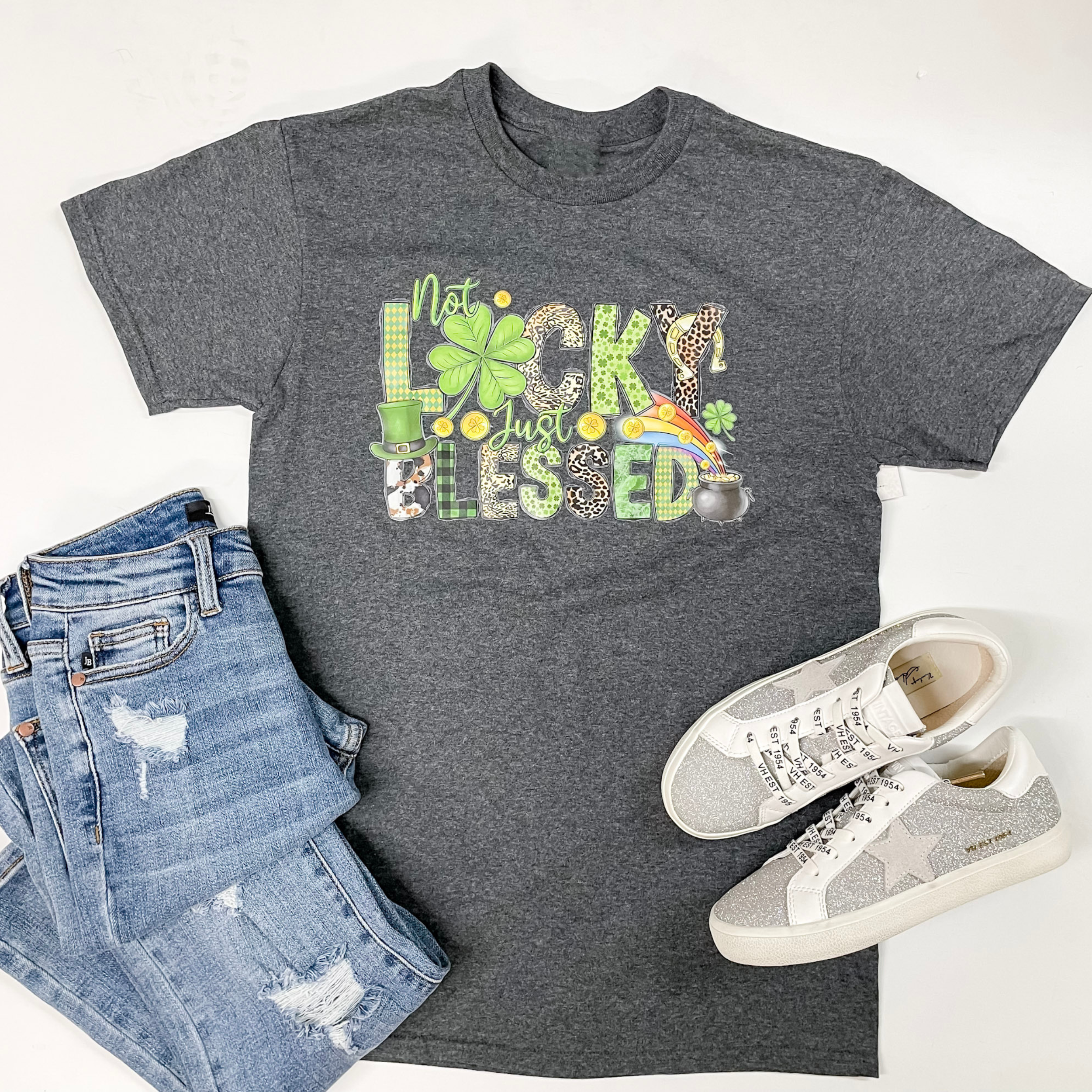 A gray tee is laid out on a white back ground. In the center of the tee are the words "Not Lucky Just Blessed." The designs in the letters vary and include designs such as plaid, cheetah, and cowprint. On the bottom left of the picture is a light-wash pair of jeans and on the right is a silver/white pair of sneakers. 