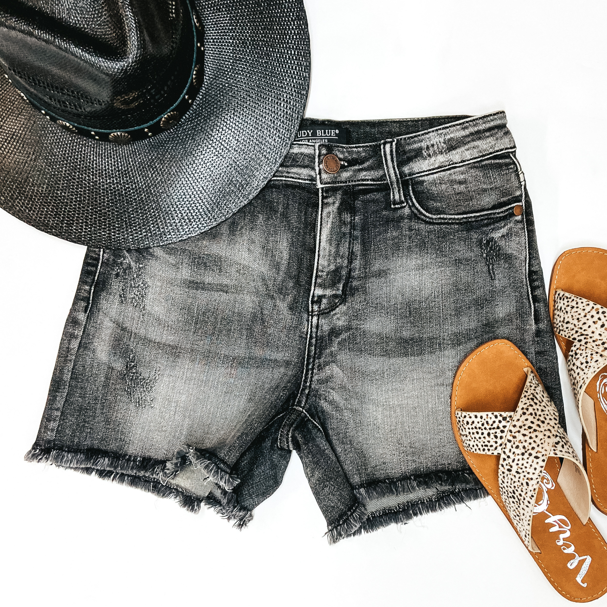 A pair of black wash denim shorts with a frayed hemline. Pictured on white background with black straw hat and dotted sandals.
