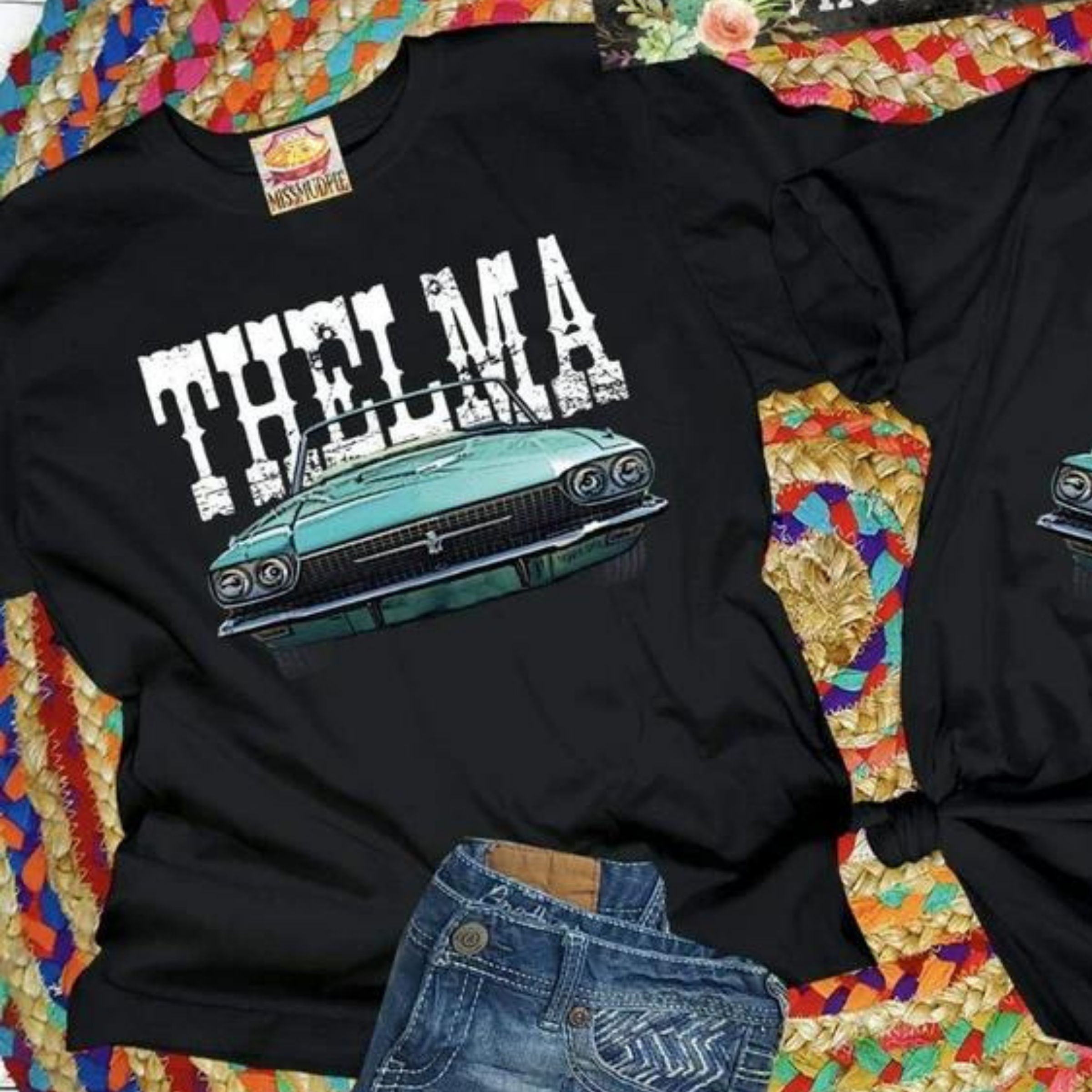A black tee shirt folded into a rectangle exposing the vintage car graphic that says "Thelma" above. Pictured with a turquoise necklace on a serape rug.