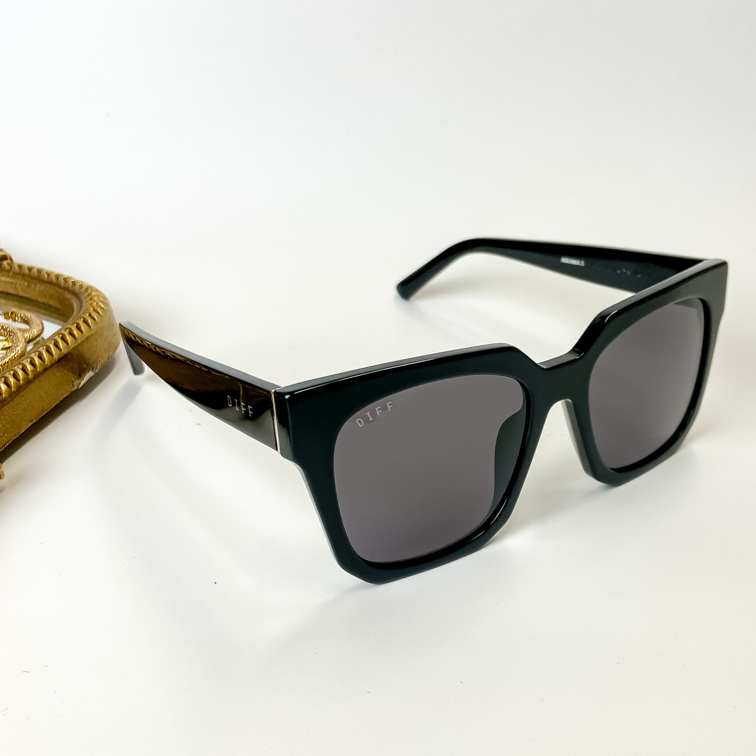 A pair of square black sunglasses with grey lenses pictured on a white background with gold jewelry.