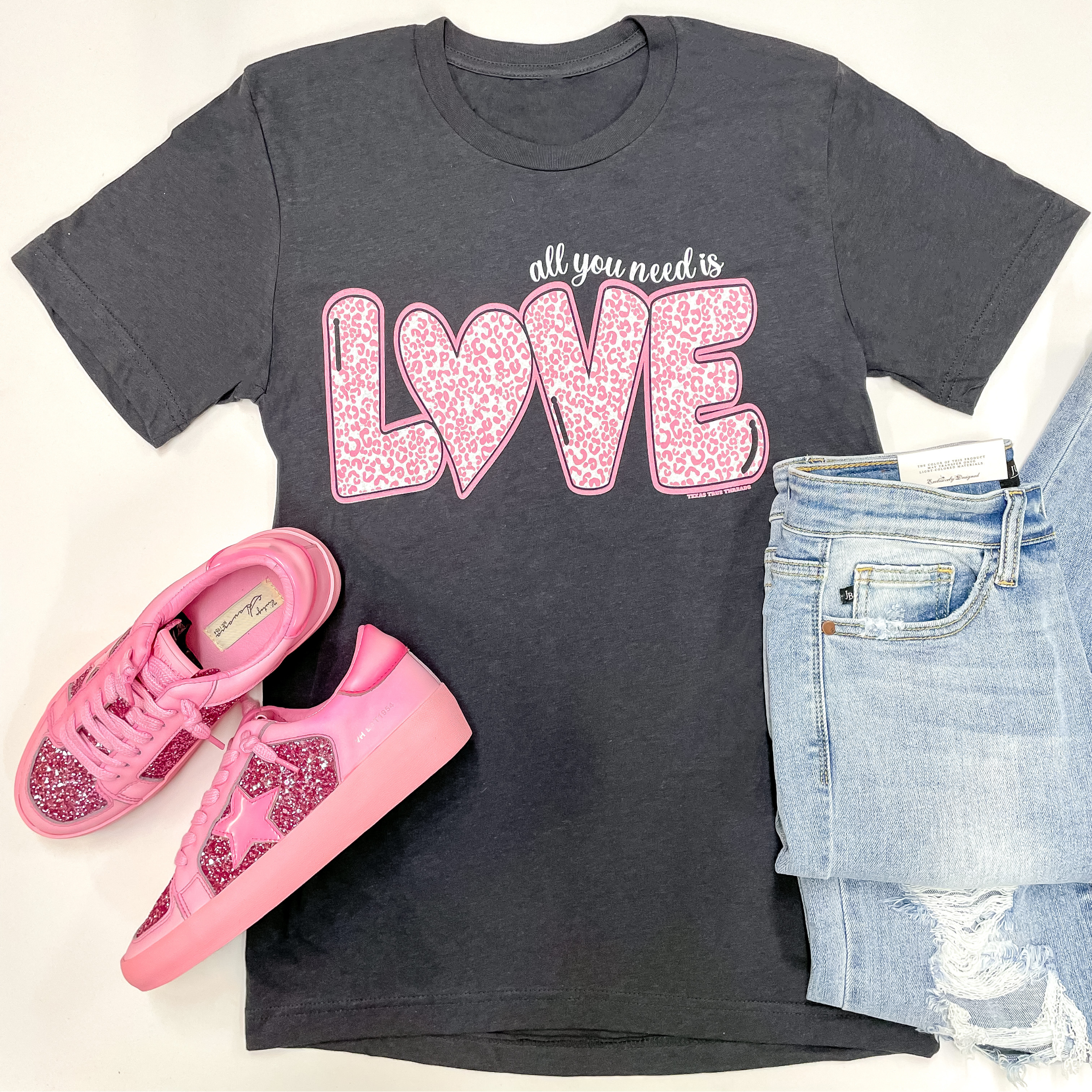 A charcoal grey short sleeve tee shirt with a graphic that says "All you need is love" in light pink leopard print. This tee is pictured on a white background with distressed jeans and hot pink sneakers to match.
