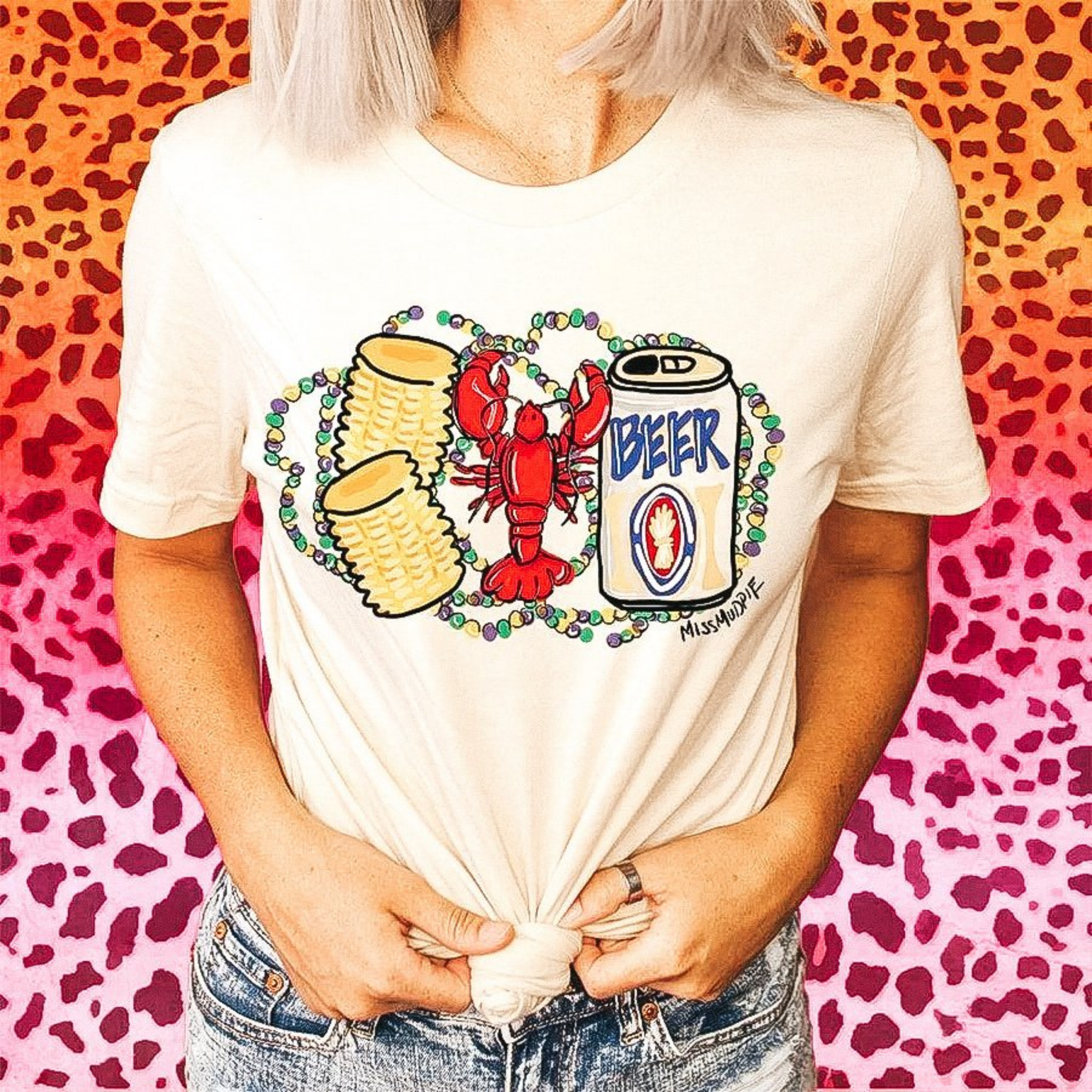 A cream colored crew neck tee shirt. The tee shirt has a graphic of corn, a crawfish, and a beer can. The graphic has mardi gras beads around it. 